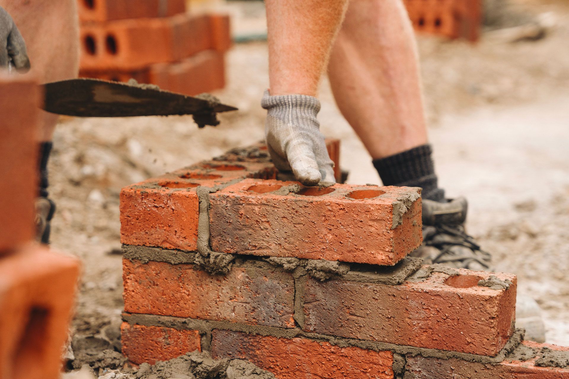 Bricklayer using a trowel to place a brick on a partially constructed brick wall; mortar visible.
