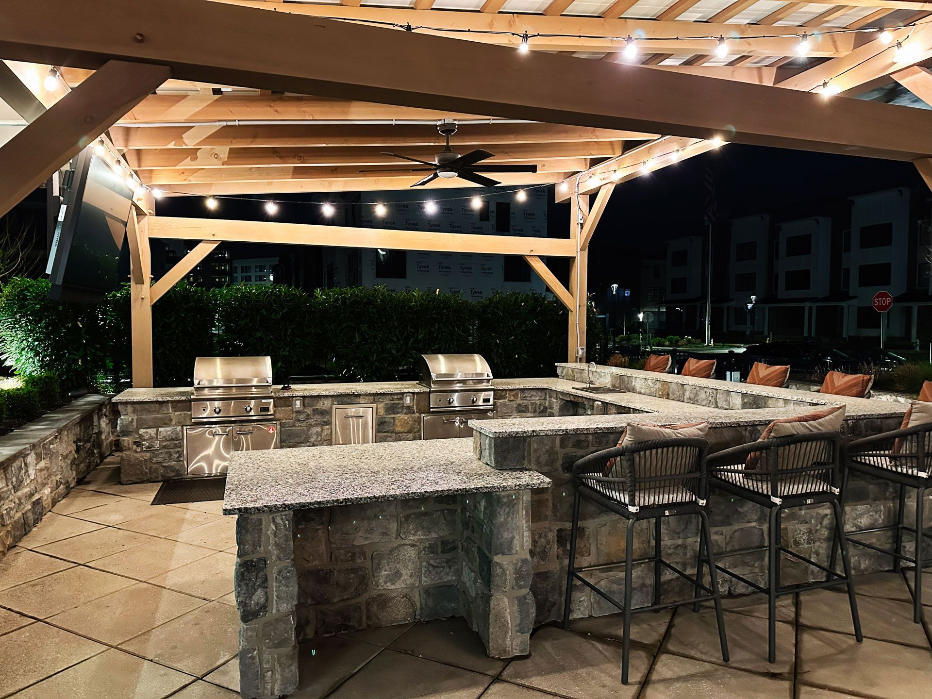 Outdoor kitchen with stone counters, grill, bar seating, and wooden pergola with string lights. Outdoor kitchen with stone counters, grill, bar seating, and wooden pergola with string lights.