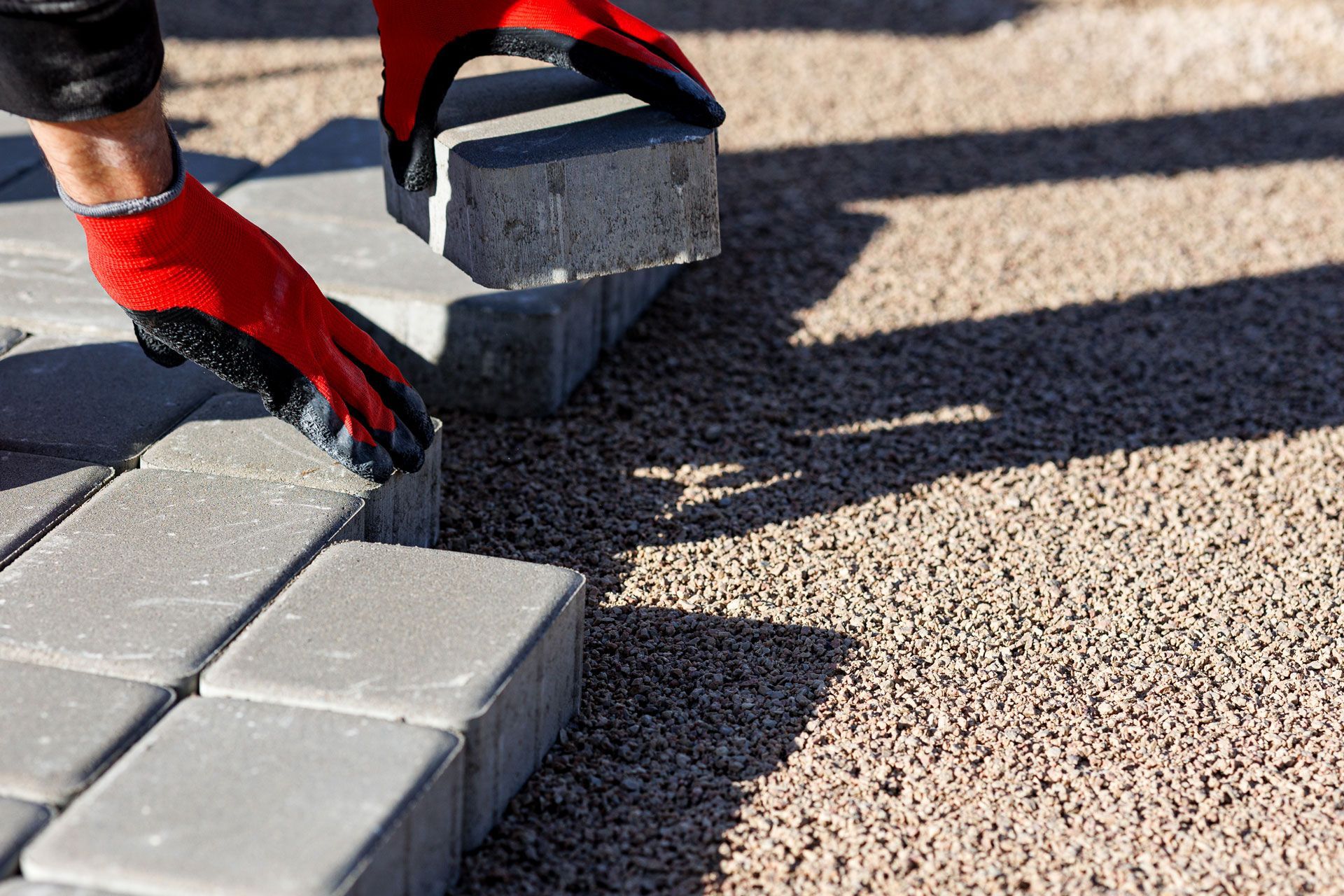 Red-gloved hands placing a gray paving stone next to other stones on a gravel surface.