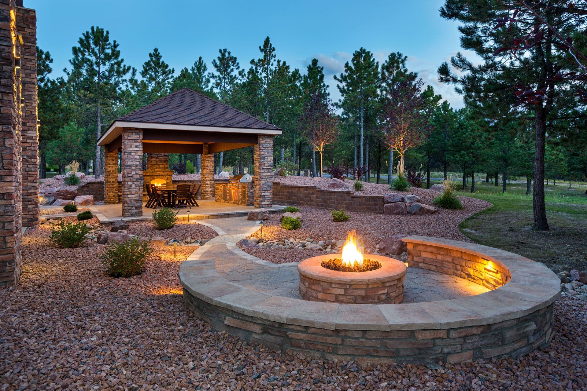 Stone patio with fire pit, seating, and gazebo surrounded by trees; evening lighting. Stone patio with fire pit, seating, and gazebo surrounded by trees; evening lighting.