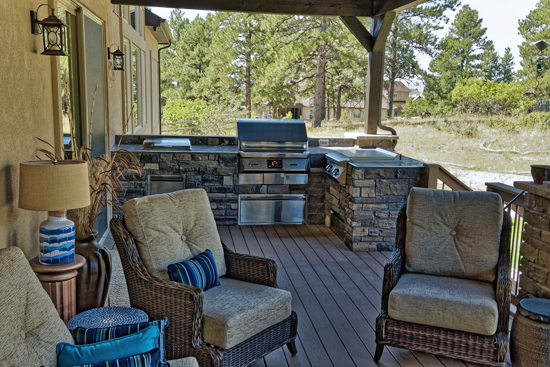 Outdoor kitchen with grill and stone countertop, wicker chairs on deck, forest backdrop.