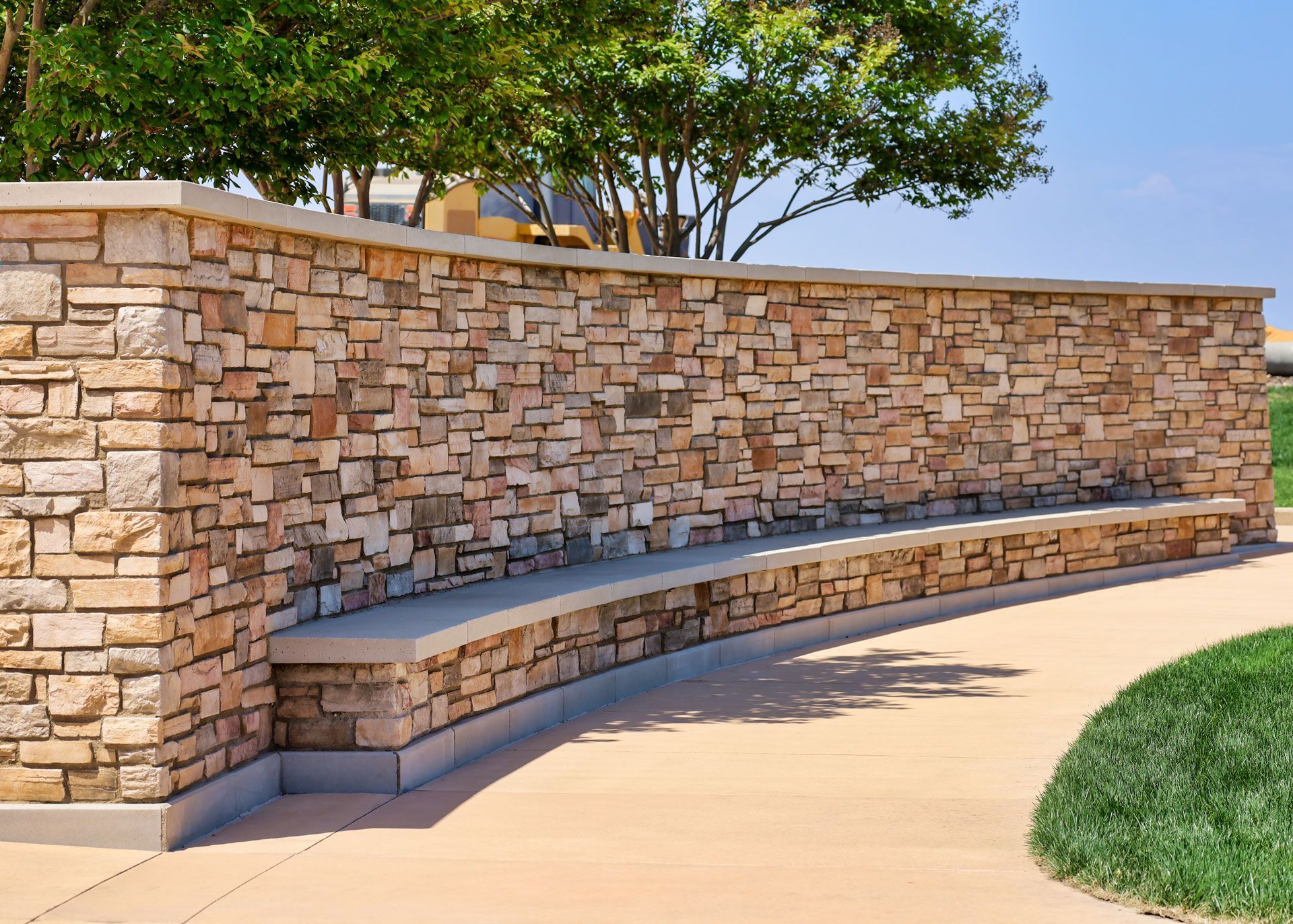 Stone wall with curved built-in bench along a paved pathway, next to a grassy area and trees. Stone wall with curved built-in bench along a paved pathway, next to a grassy area and trees.