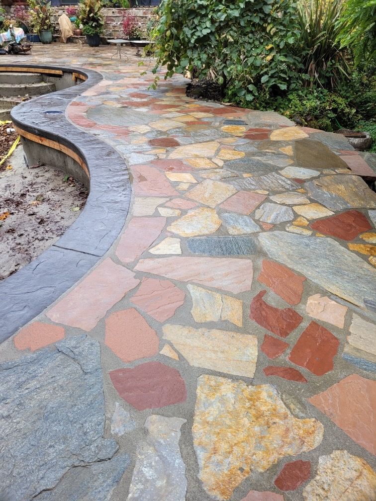 A partially constructed stone fire pit and retaining wall on a stone patio in a backyard setting.