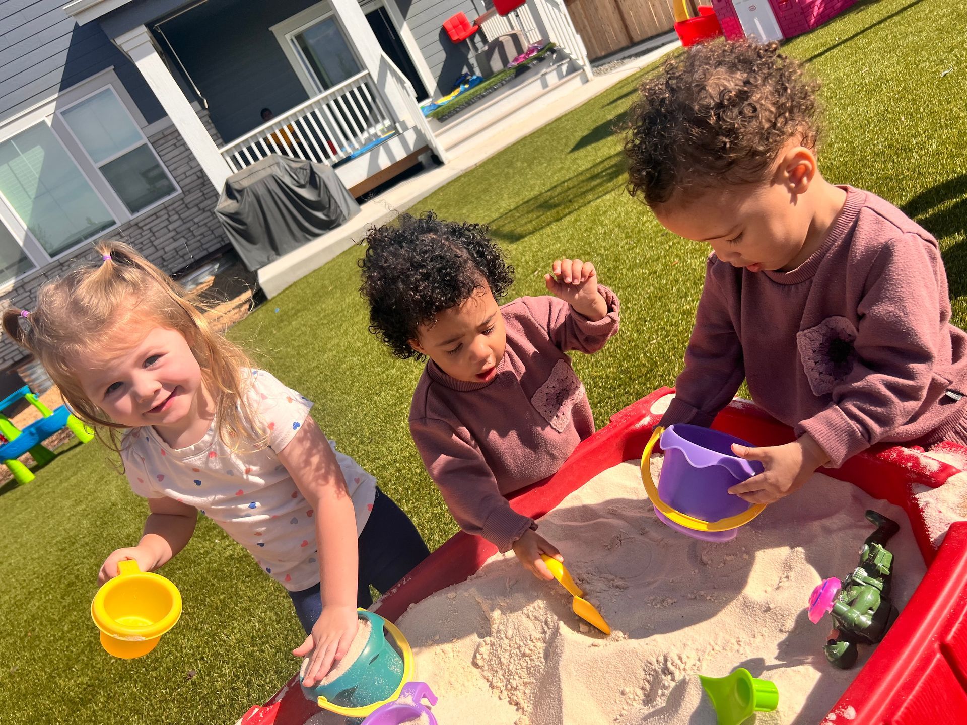 Children playing with sand in a red sandbox on green turf; sunny outdoor setting.