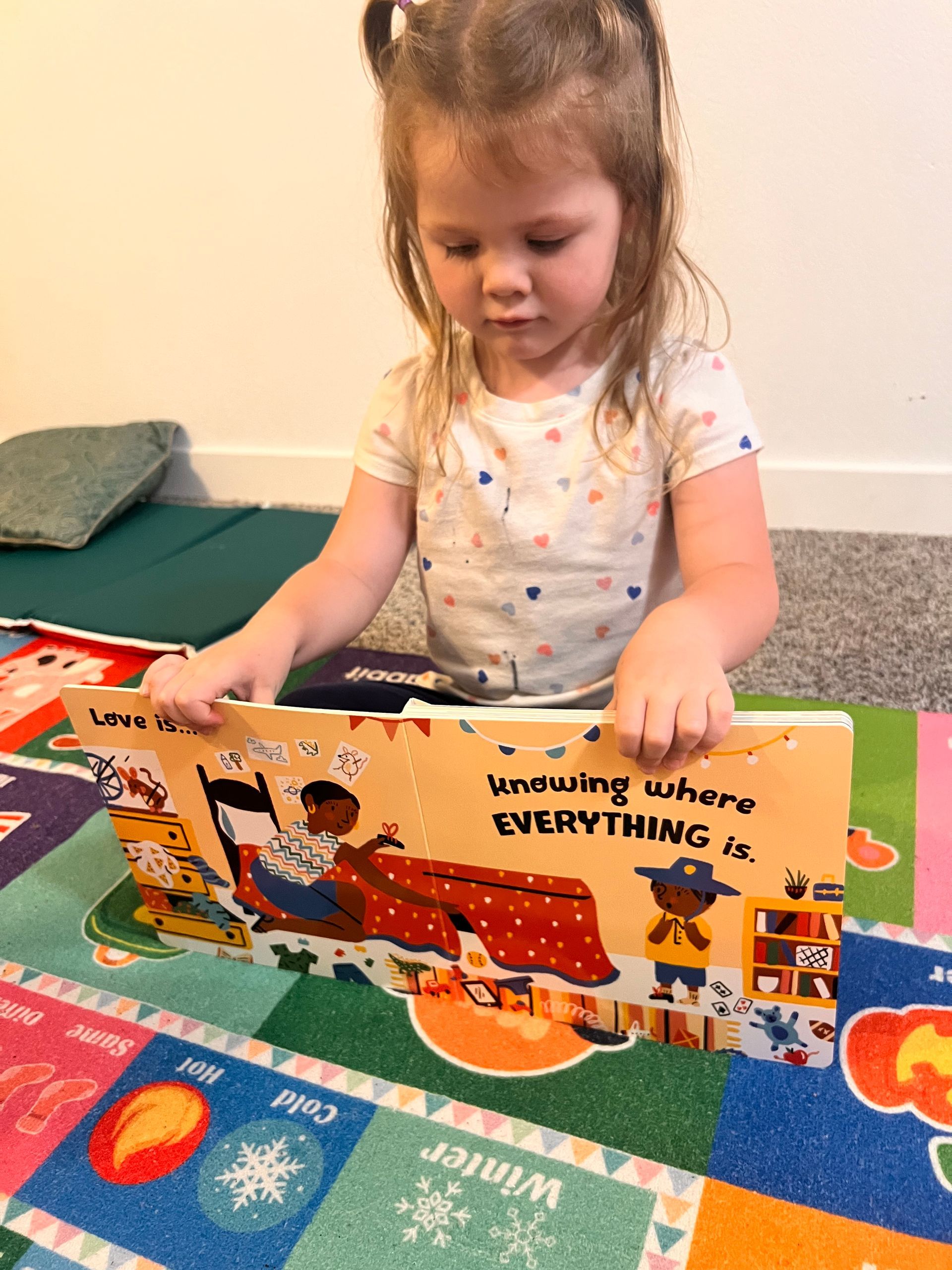 Young child reading a book on a colorful rug. The book shows illustrations and text.