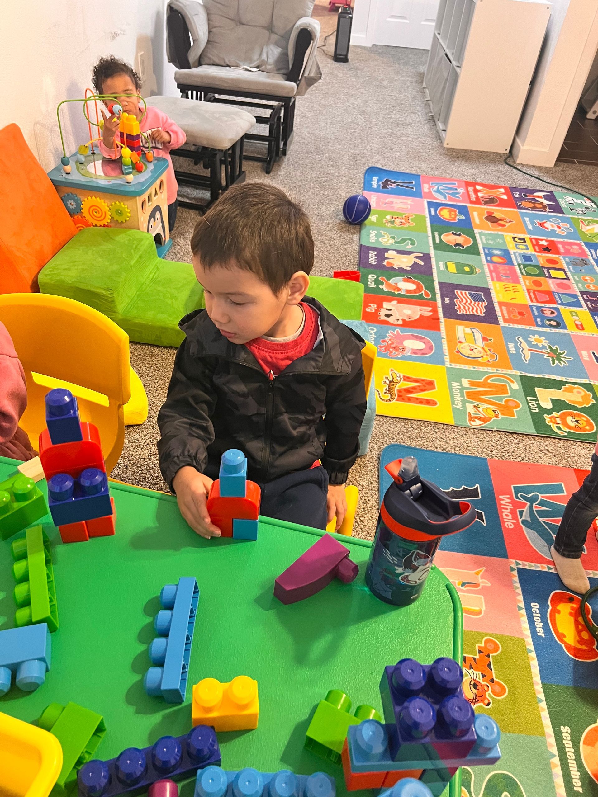 Boy playing with building blocks at a table, another child in the background. Colorful play area with toys.