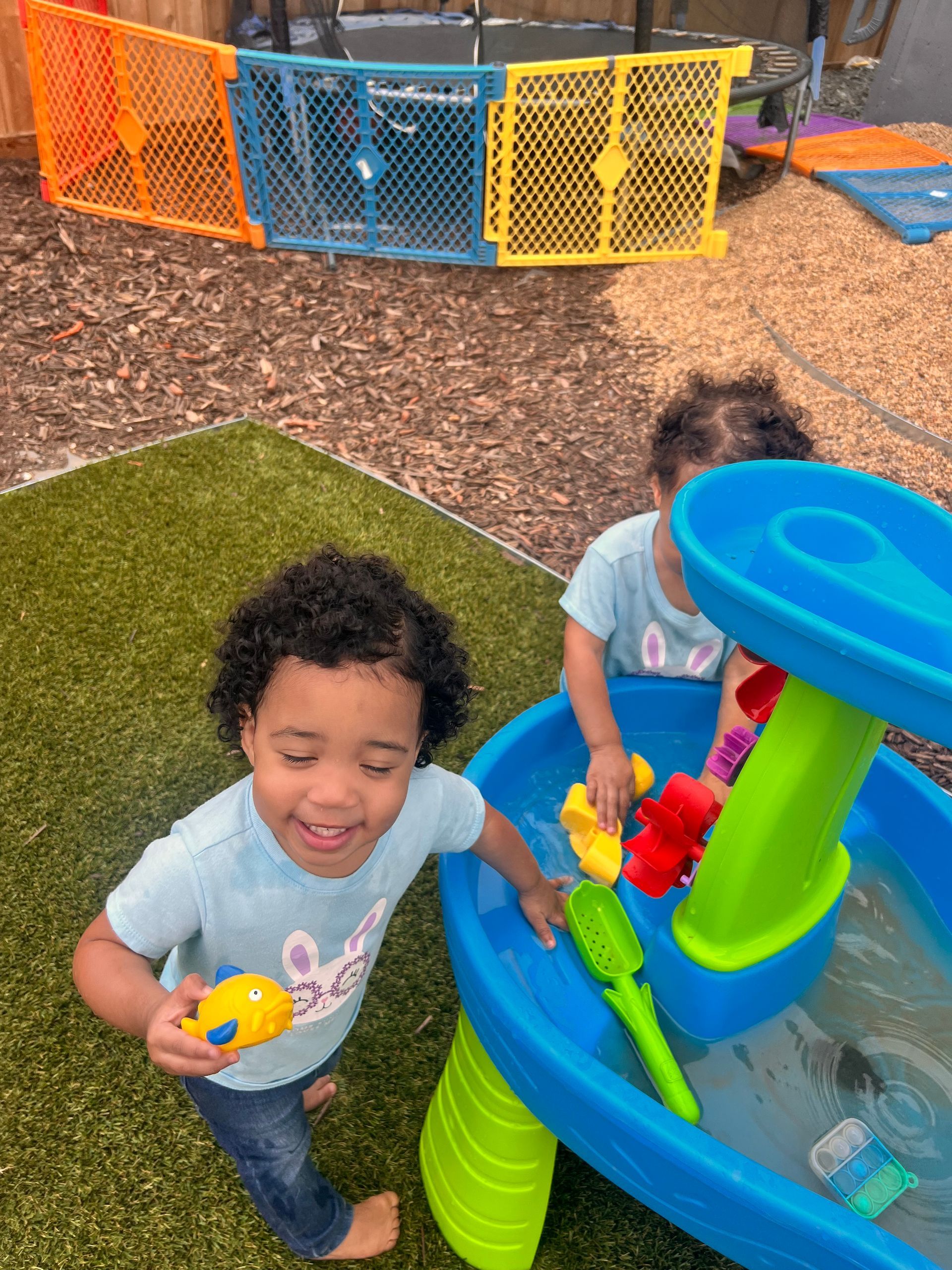 Two children play at a water table outdoors, one holding a toy, smiling. Orange, blue, yellow fence in background.