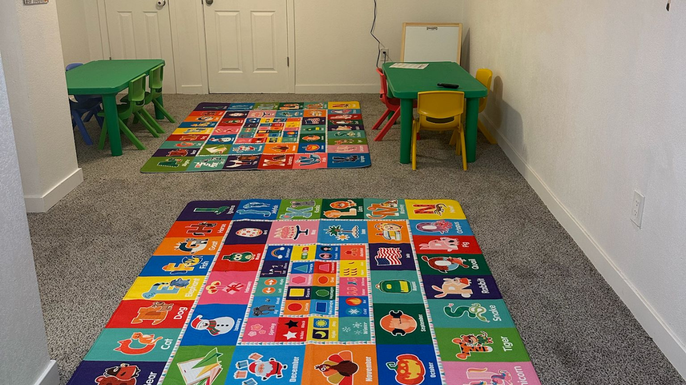 Playroom with colorful alphabet rugs, green tables, and yellow chairs on gray carpet.