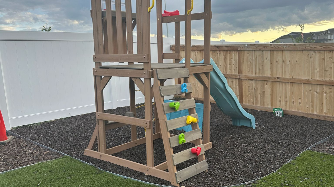 Play set with climbing wall and slide in a backyard with black mulch.