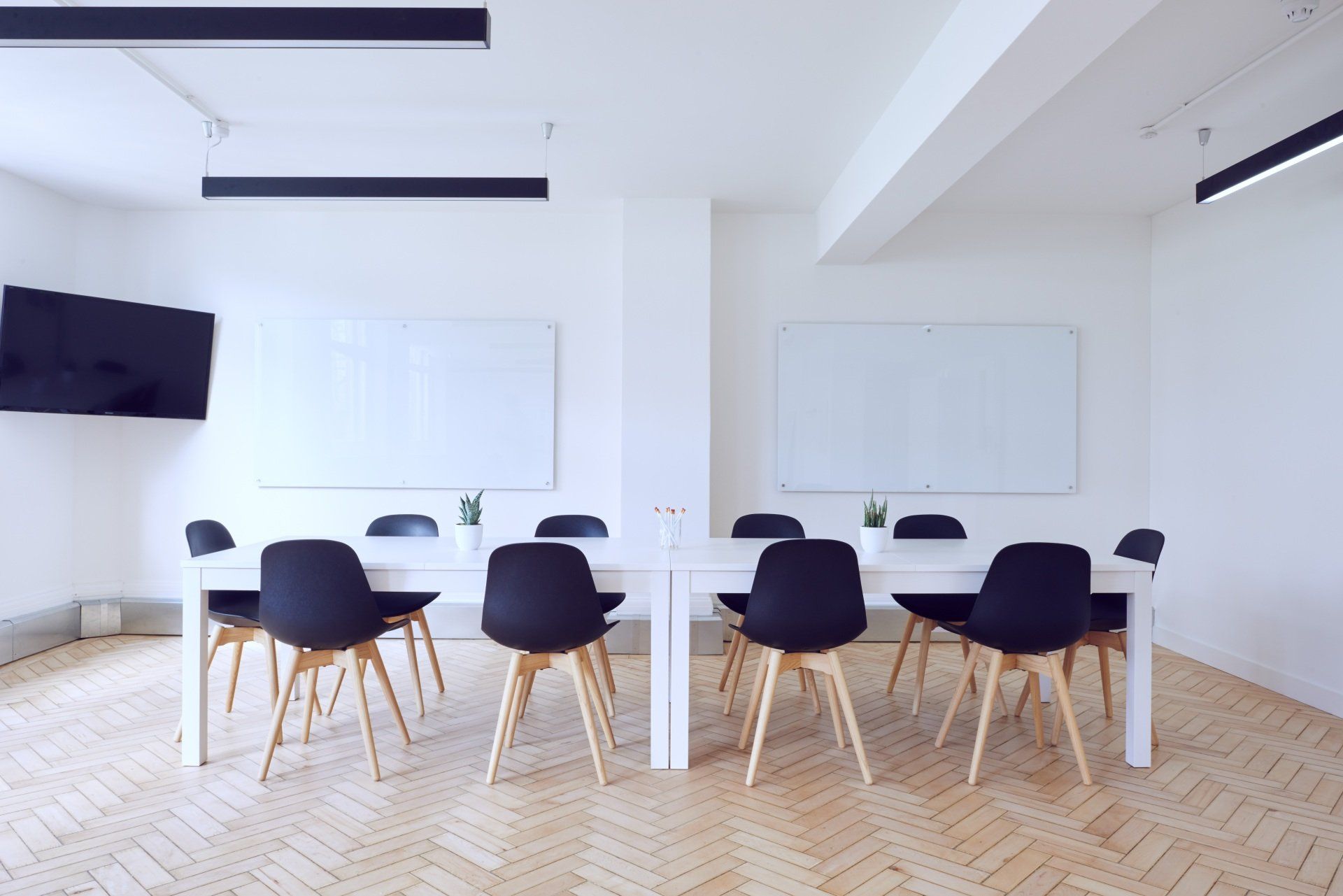 White meeting room with long table, black chairs, whiteboards, and a mounted TV.