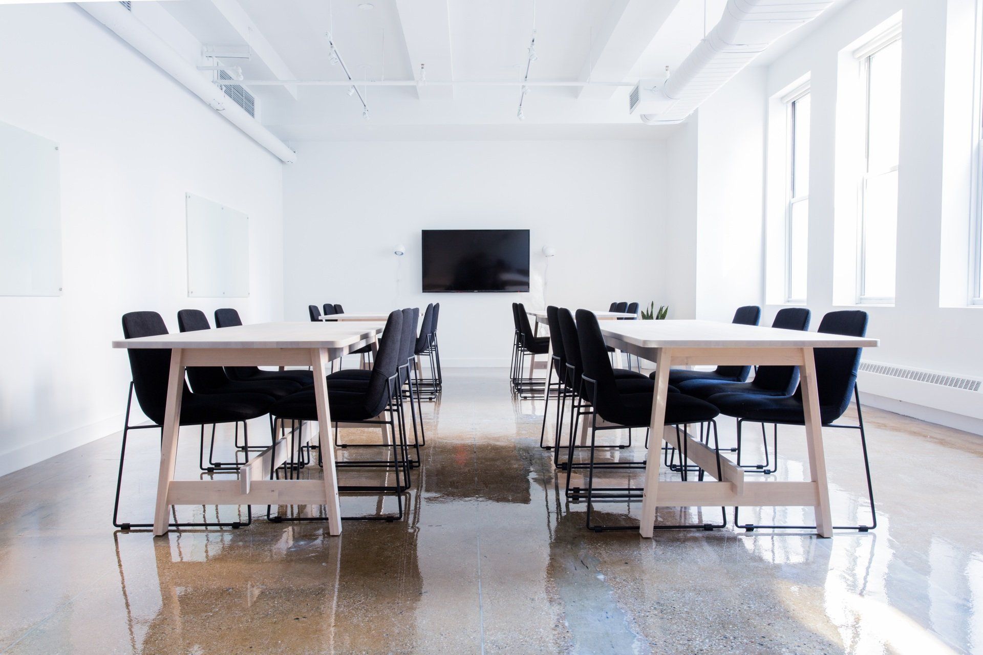 Bright, empty classroom with light tables, black chairs, and a large screen on a white wall.