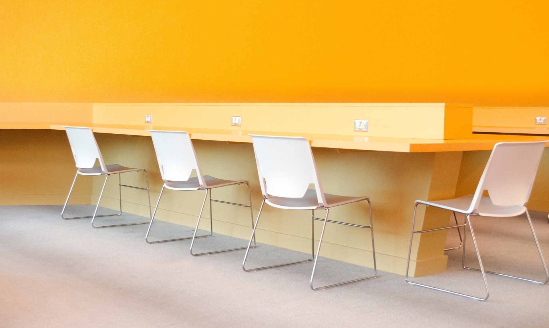 Row of white chairs at a long wooden counter in a bright orange room.