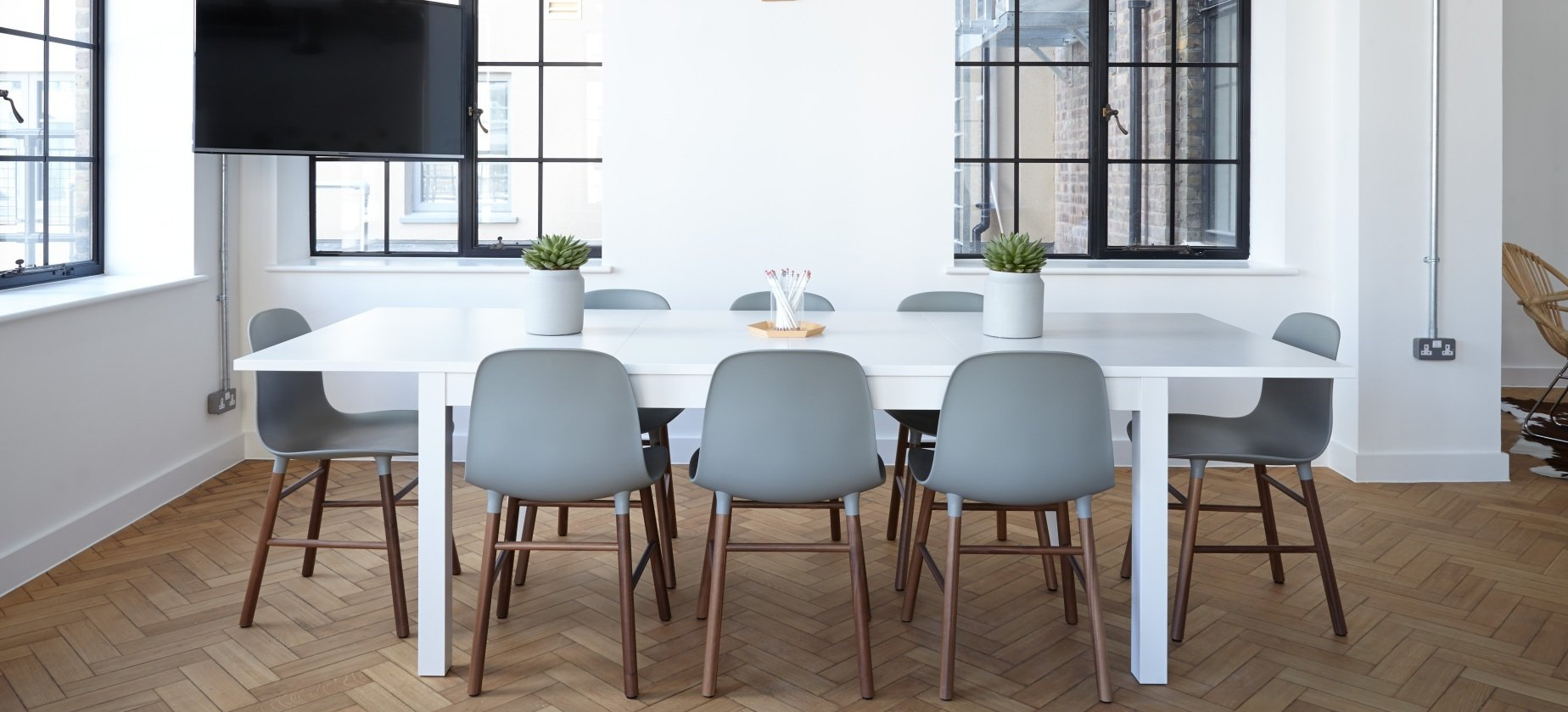 A modern office conference room with a long white table, gray chairs, and windows.