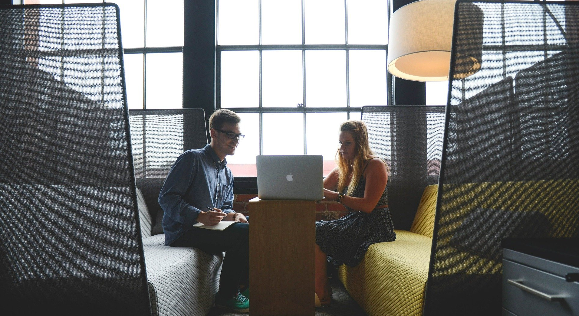 Two people sit, working together, near a window, using a laptop, and discussing paperwork.