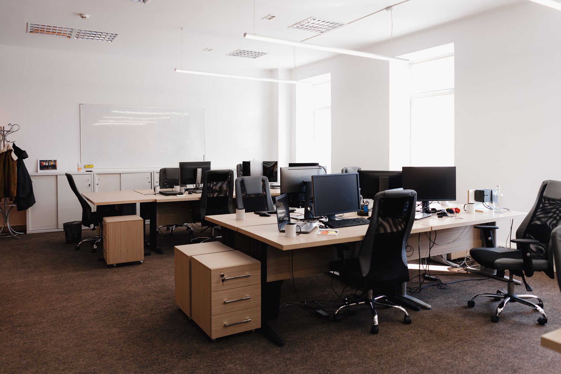 Modern office with rows of wooden desks, computer monitors, and black office chairs on a brown carpeted floor.