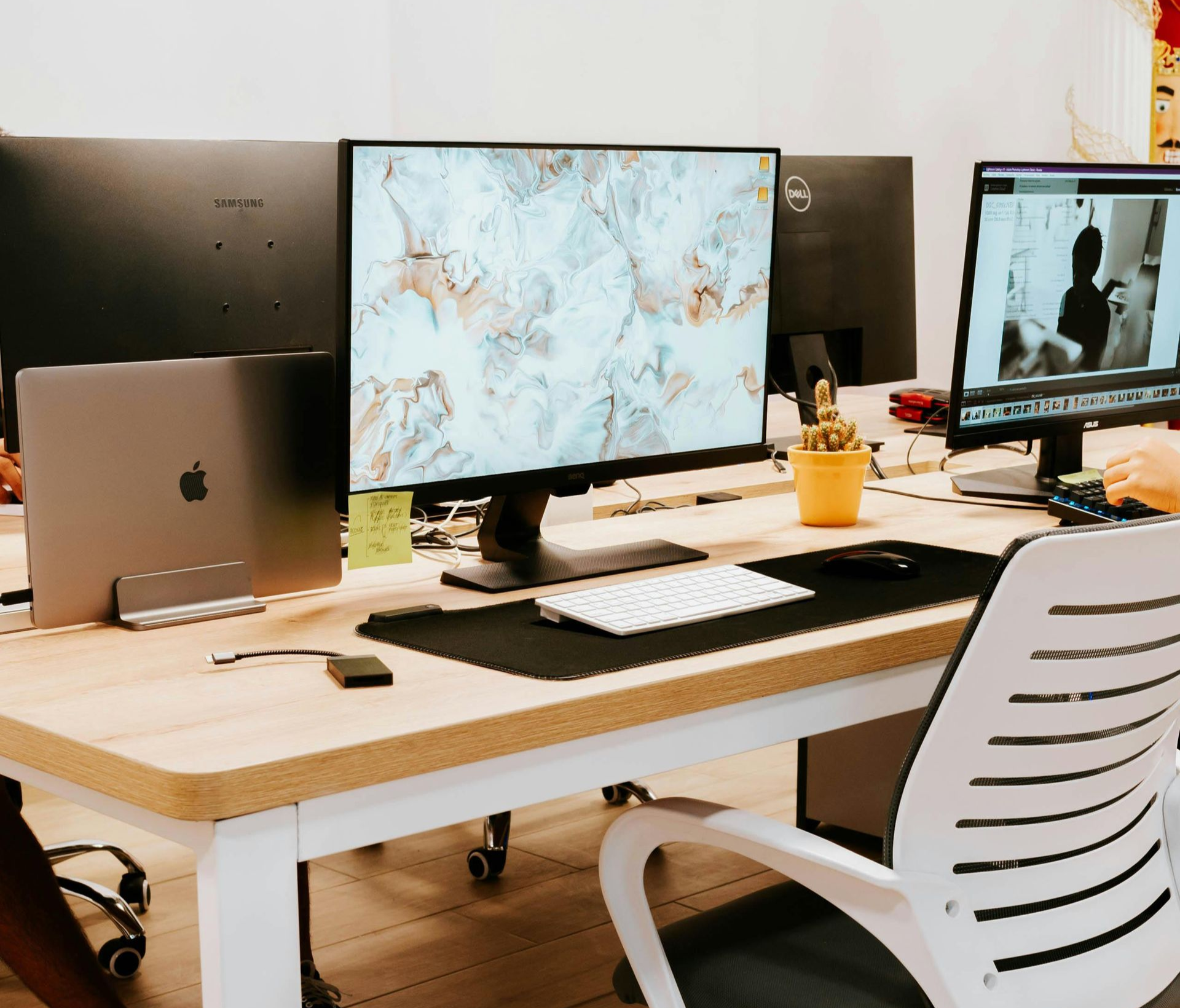 A modern, light-wood desk with a closed laptop, a monitor displaying a marbled background, and a small potted cactus.
