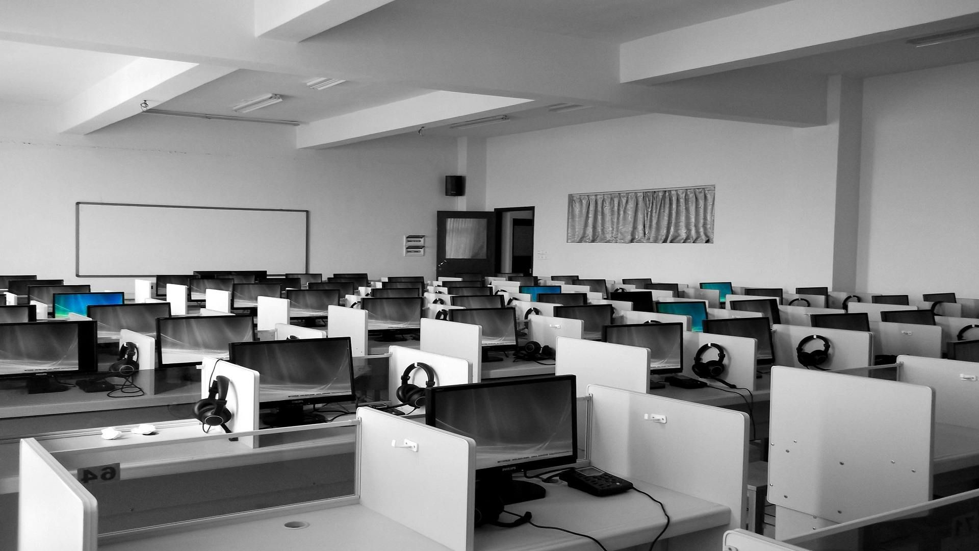 A classroom or computer lab filled with rows of desks, monitors, and headphones under bright lighting.