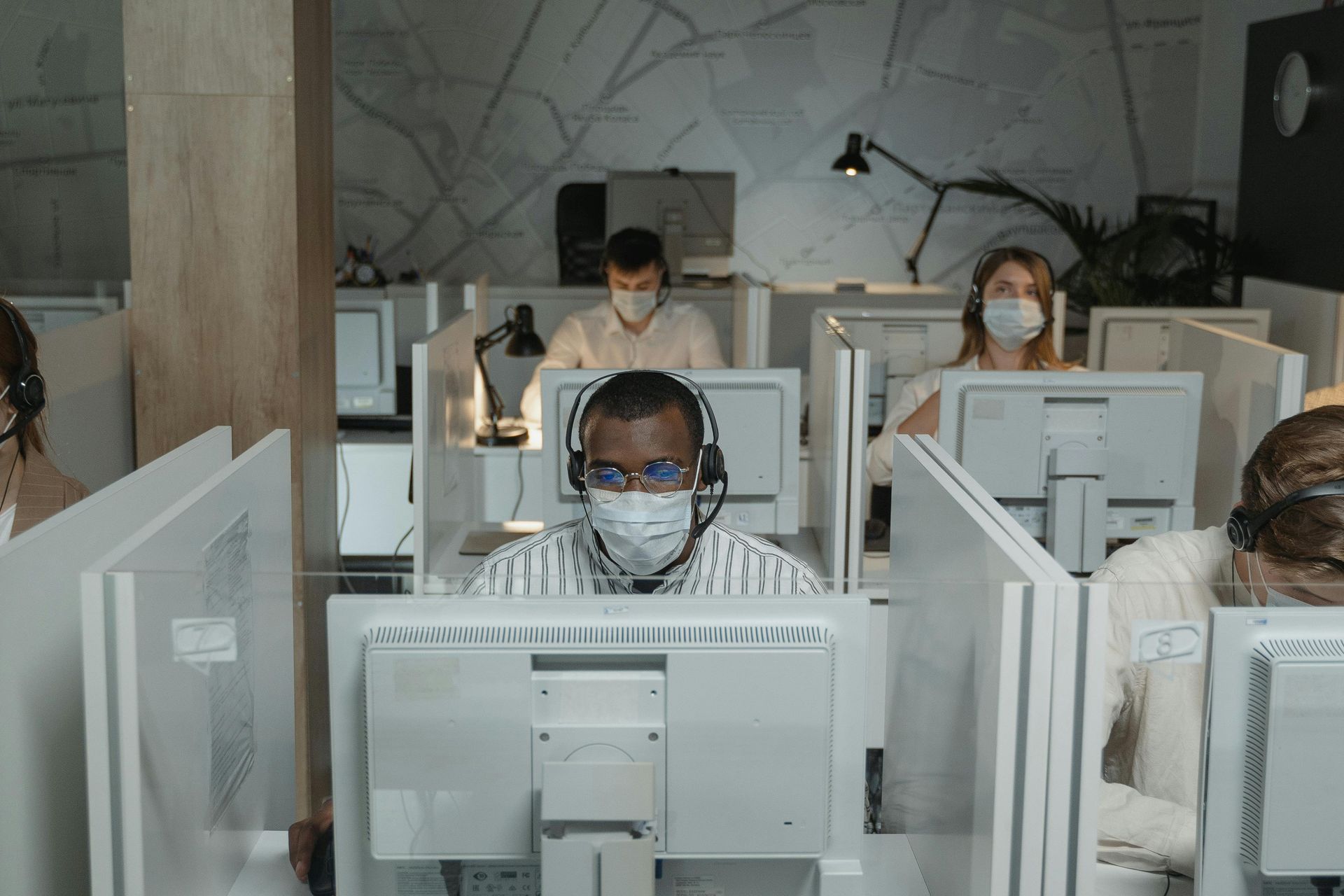 A group of employees wearing headsets and masks working at computers in an office with cubicle dividers.