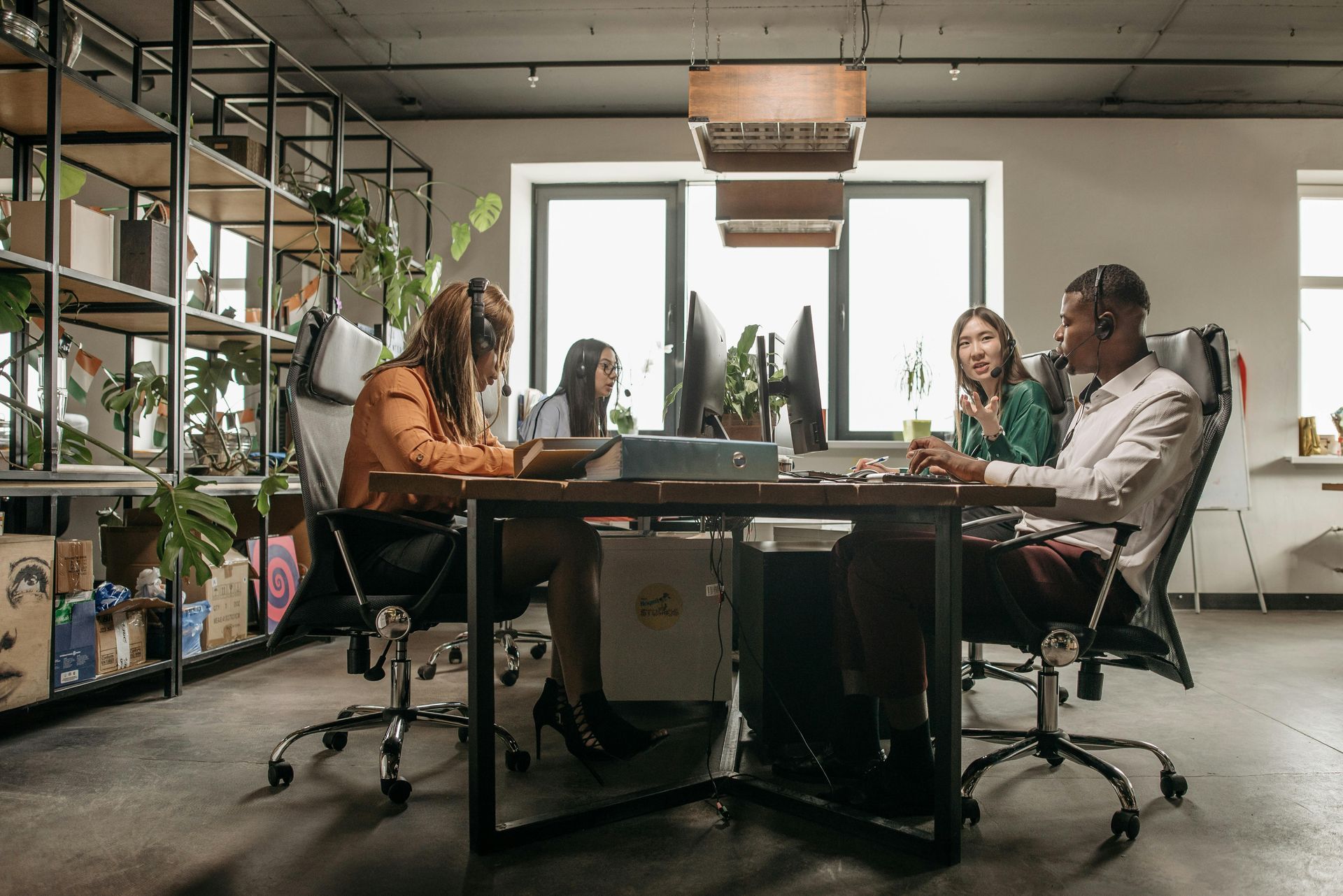 Four colleagues work at a large wooden desk in a modern, plant-filled office with industrial shelving and metal chairs.