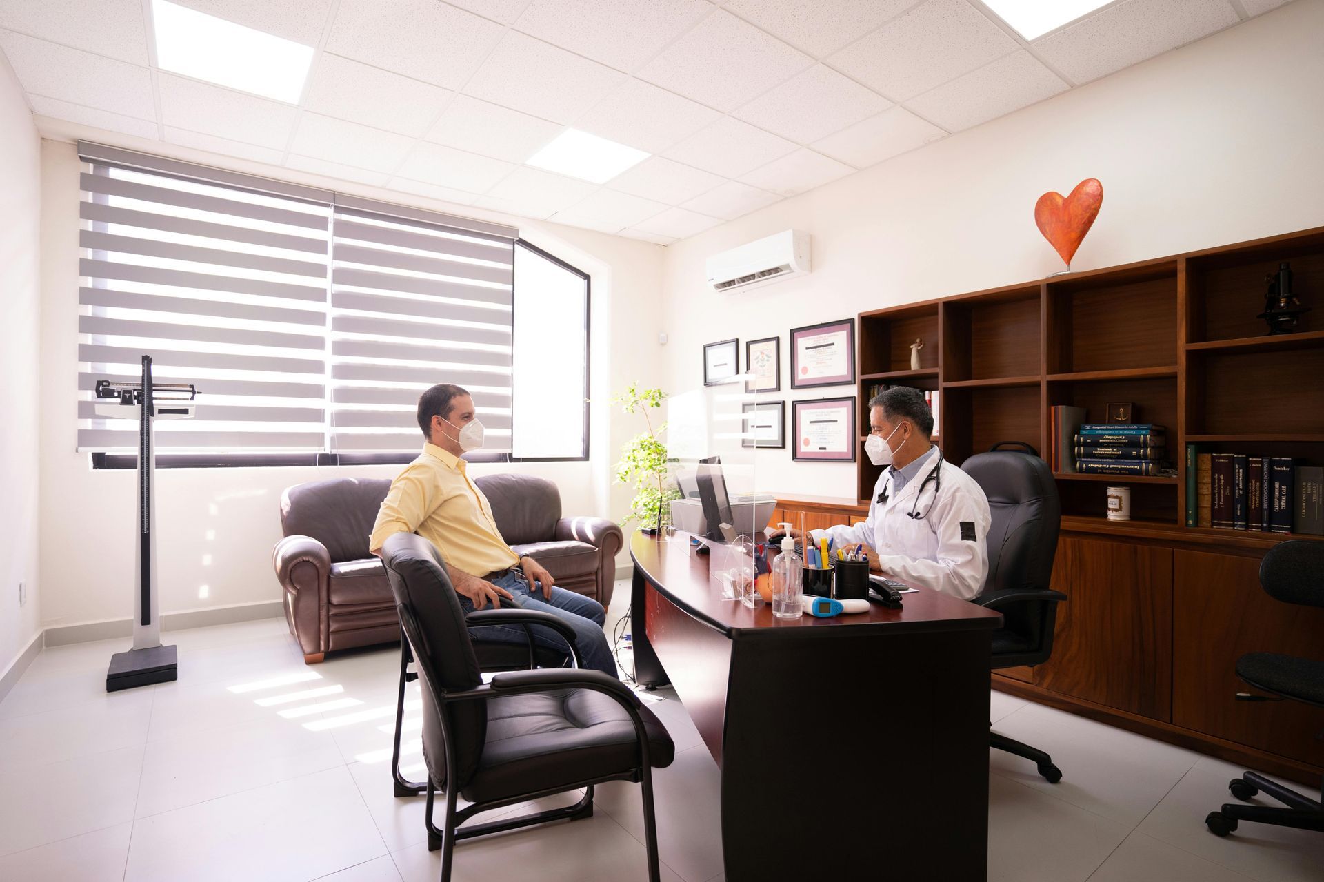 A masked doctor sits at a wooden desk in an office, speaking with a masked patient sitting in a chair across from him.