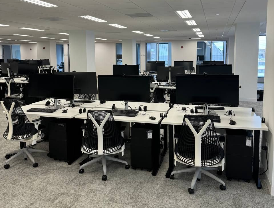 Rows of empty computer workstations in a modern office, featuring black desktops, white desks, and ergonomic chairs.