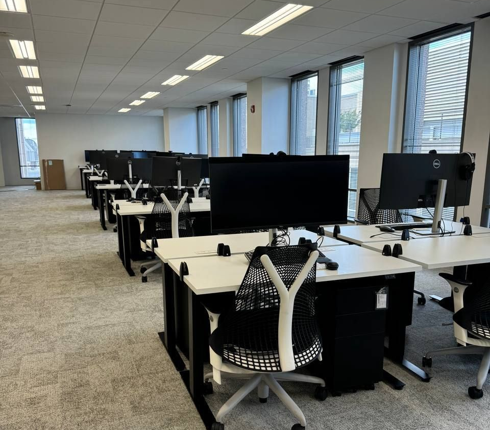 Empty office space with rows of desks, black chairs, and large computer monitors, under fluorescent lights.