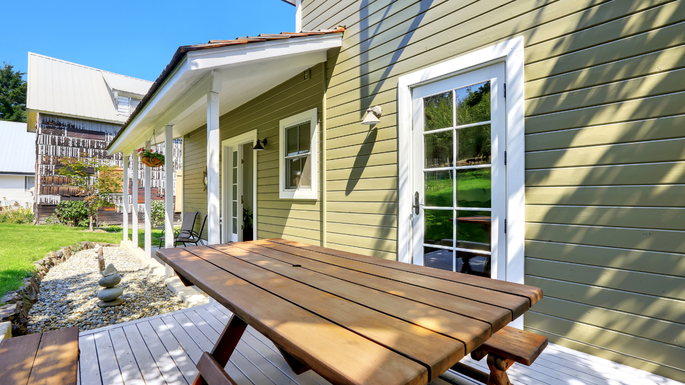 A wooden picnic table is on a deck in front of a house.