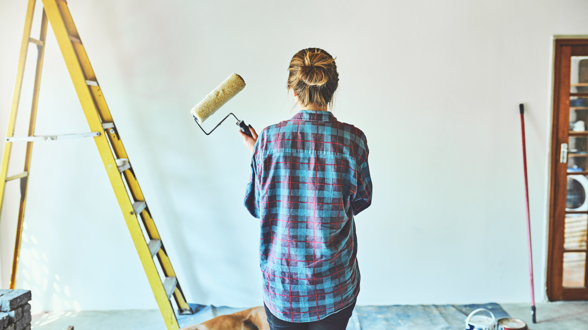 Woman painting a white wall with a paint roller; near a ladder, in a room.