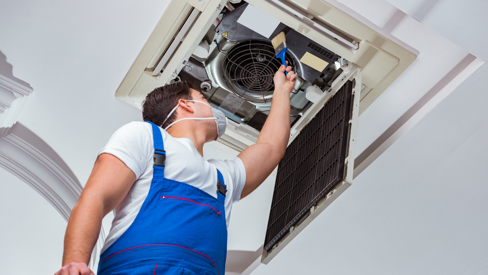 Man in blue coveralls cleaning a ceiling air conditioning unit with a brush, wearing a mask.