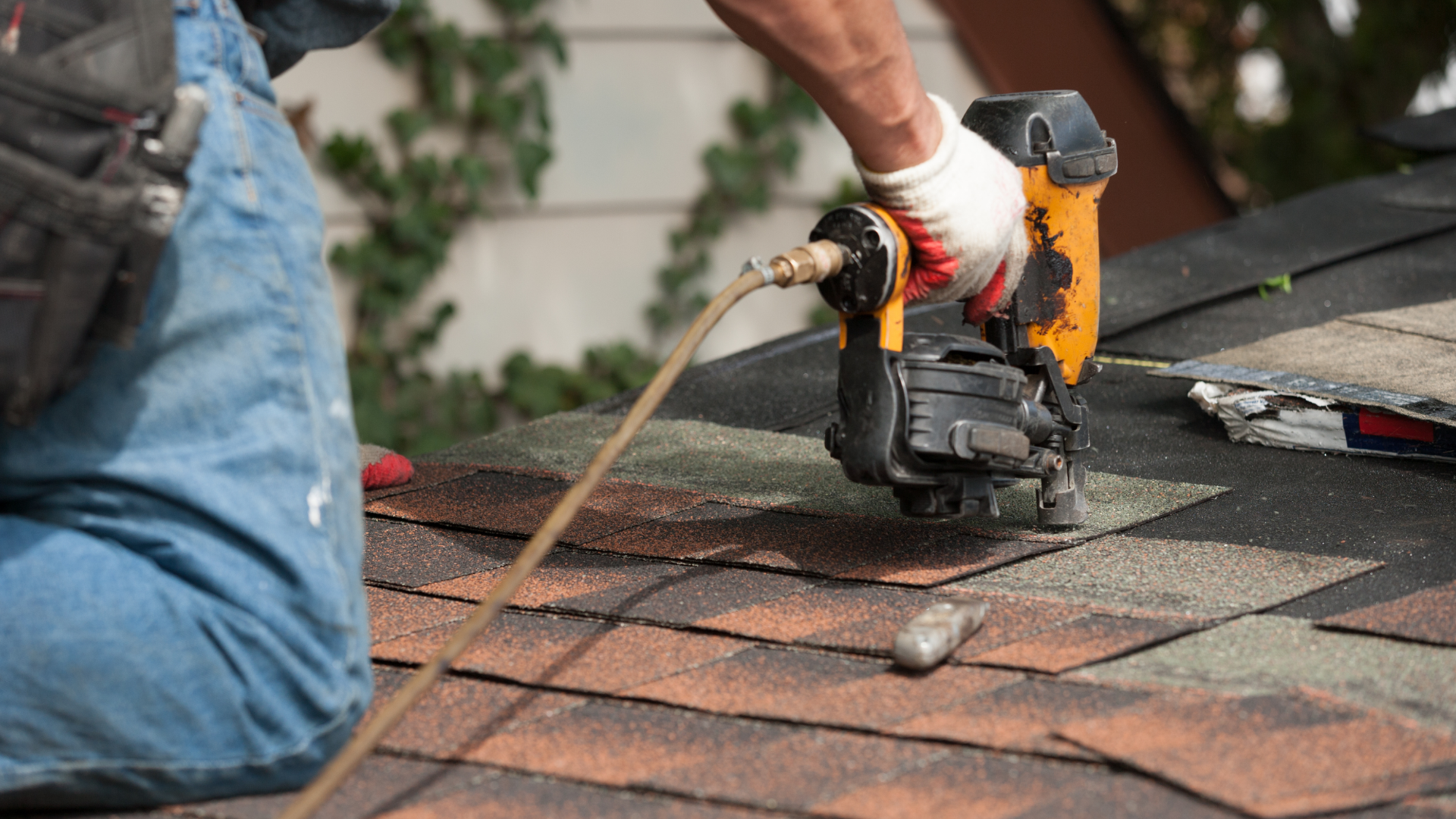 Roofer using a nail gun on a shingle roof; tan, brown, and grey shingles.