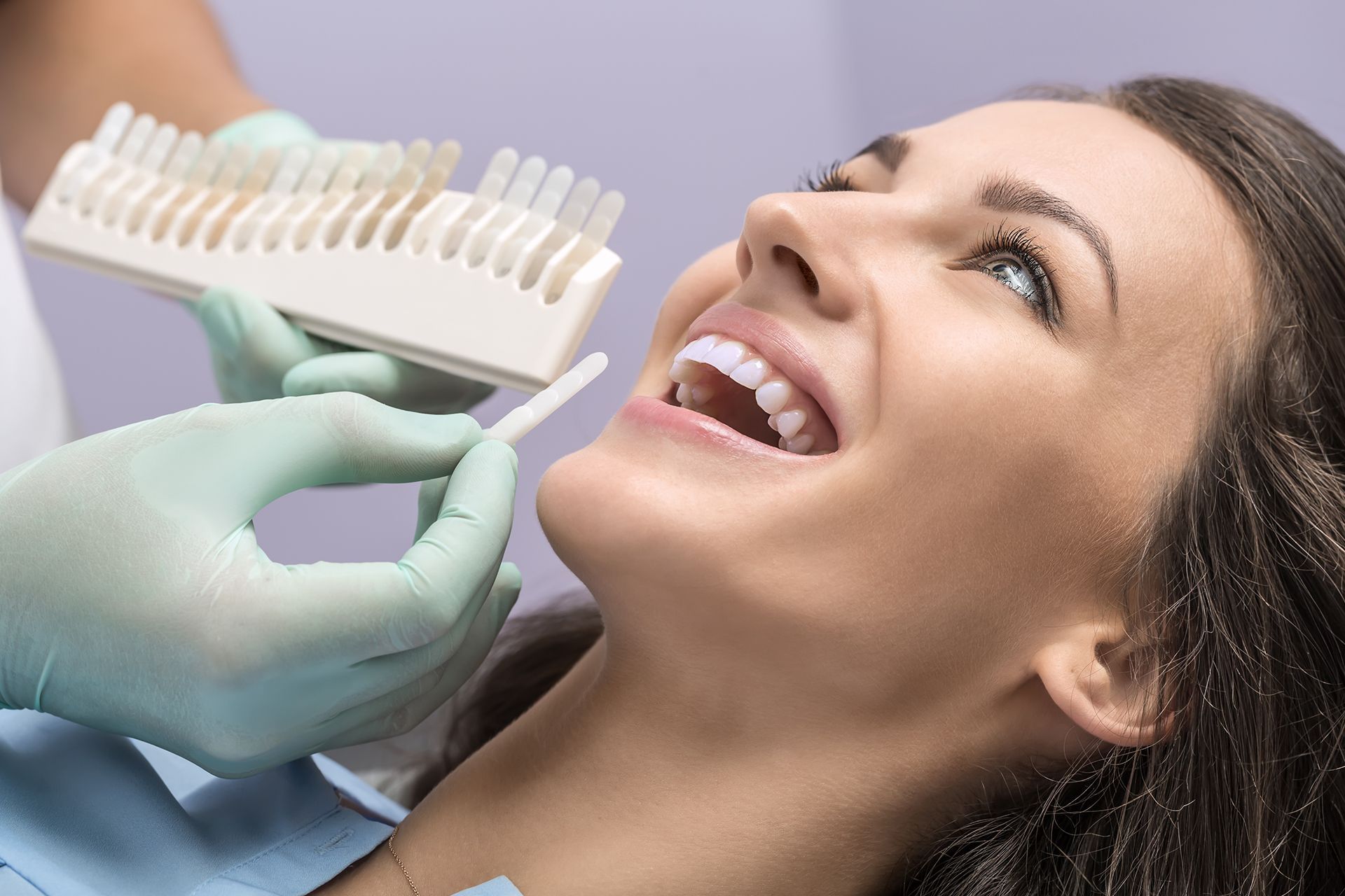 Dentist using a shade guide to assess a patient's teeth color. Patient smiles, dentist wears gloves. Dentist using a shade guide to assess a patient's teeth color. Patient smiles, dentist wears gloves.