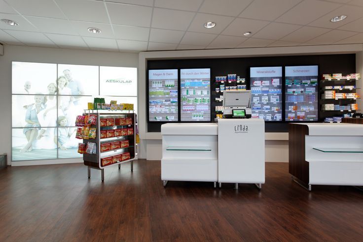 Innenansicht einer Apotheke mit weißen Theken, Regalen voller Produkte und beleuchteten Auslagen an einer dunklen Wand.