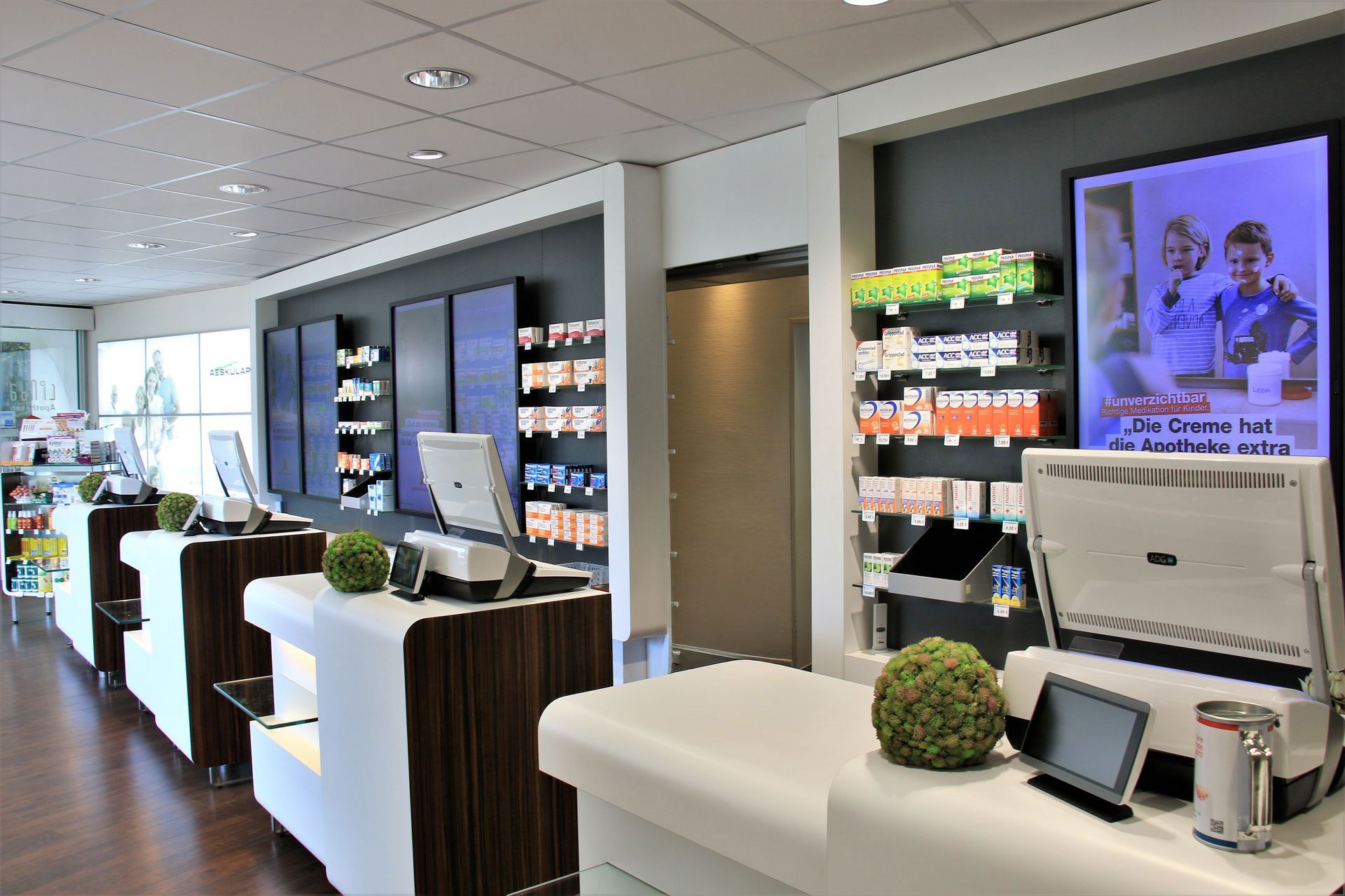 Innenansicht einer modernen Apotheke mit weißen Theken, dunklen Holzelementen und Regalen voller Medikamente.