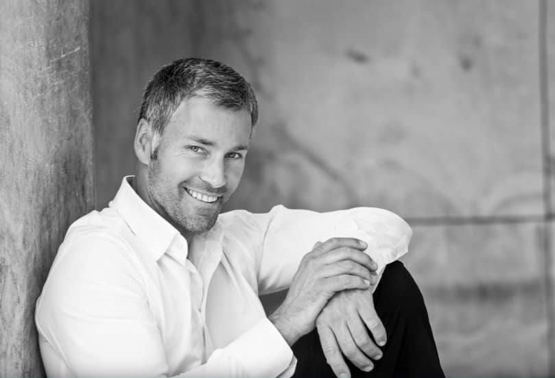 A man in a white shirt is leaning against a wall and smiling.