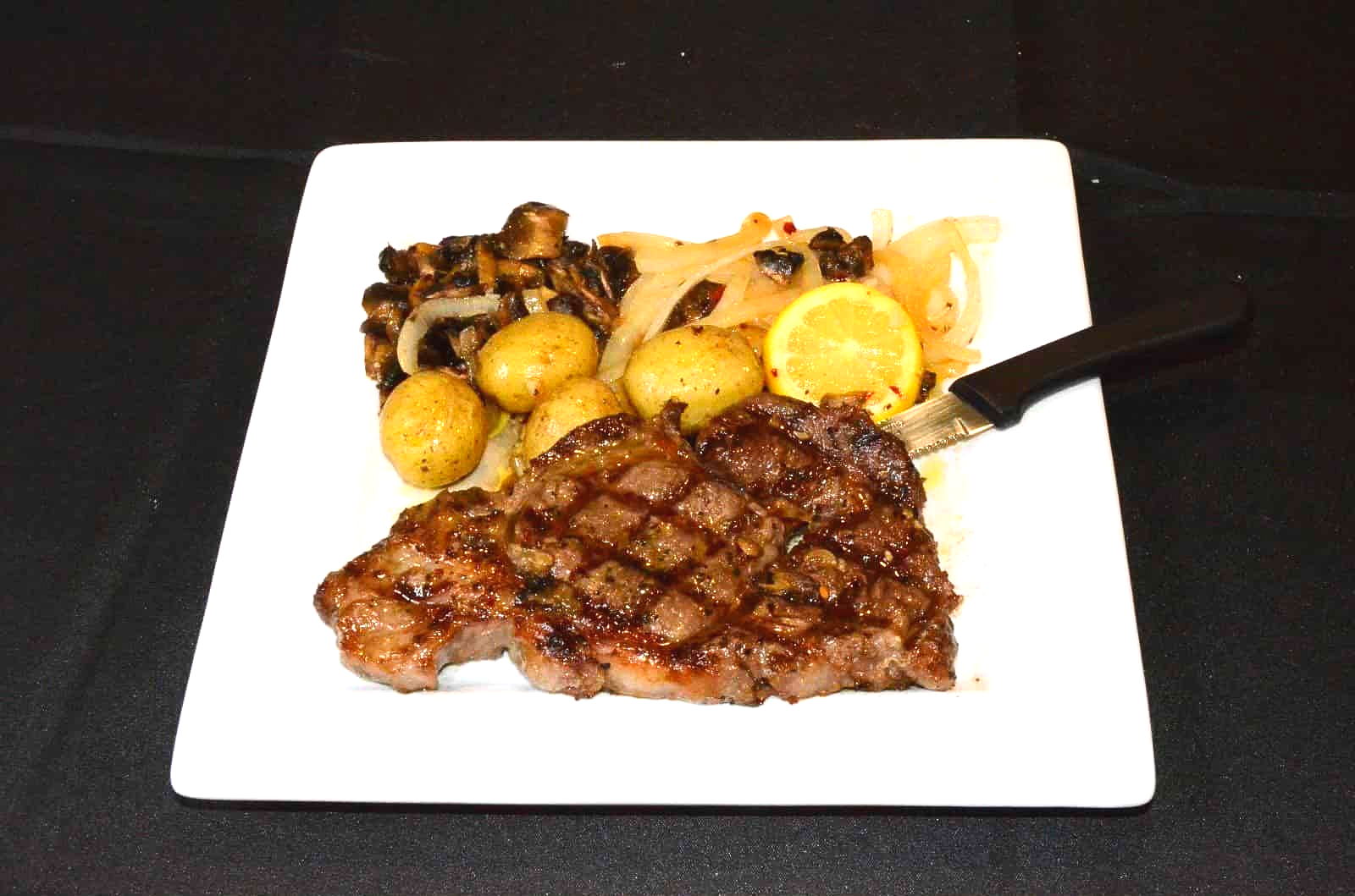 Grilled steak, potatoes, sautéed onions and lemon on a white plate.