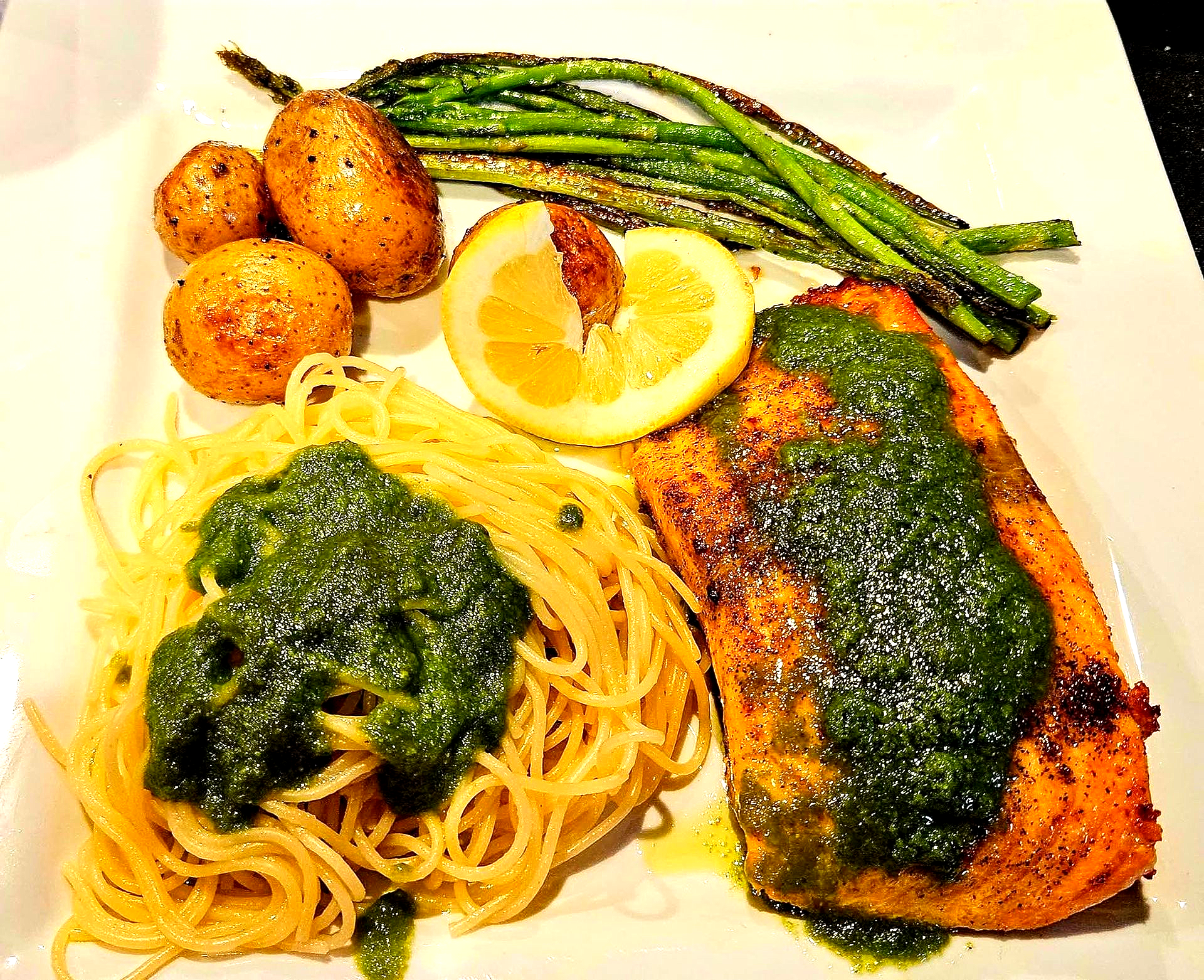 Salmon with pesto, spaghetti, roasted potatoes, and asparagus on a white plate, garnished with lemon.
