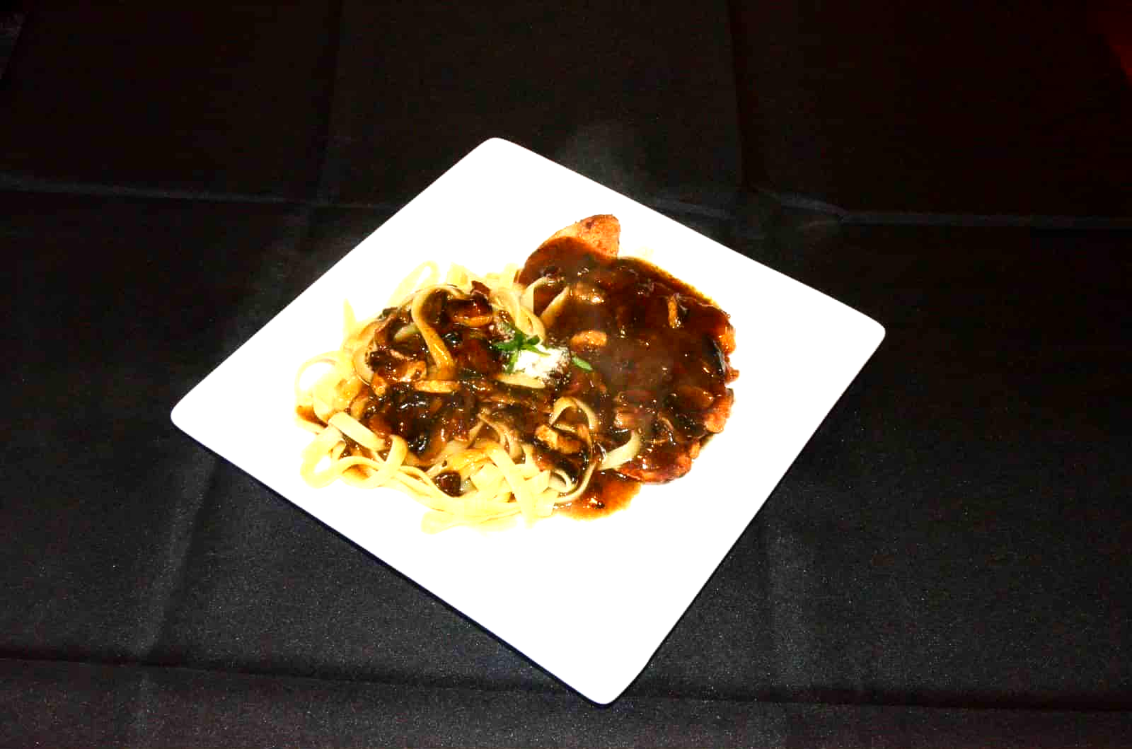 Steak with dark sauce and noodles on a white square plate, against a black background.