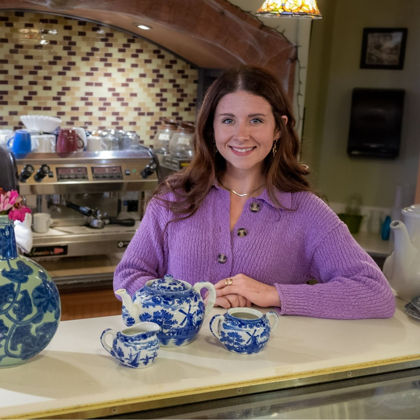 A woman in a purple sweater is sitting at a counter with a teapot and cups