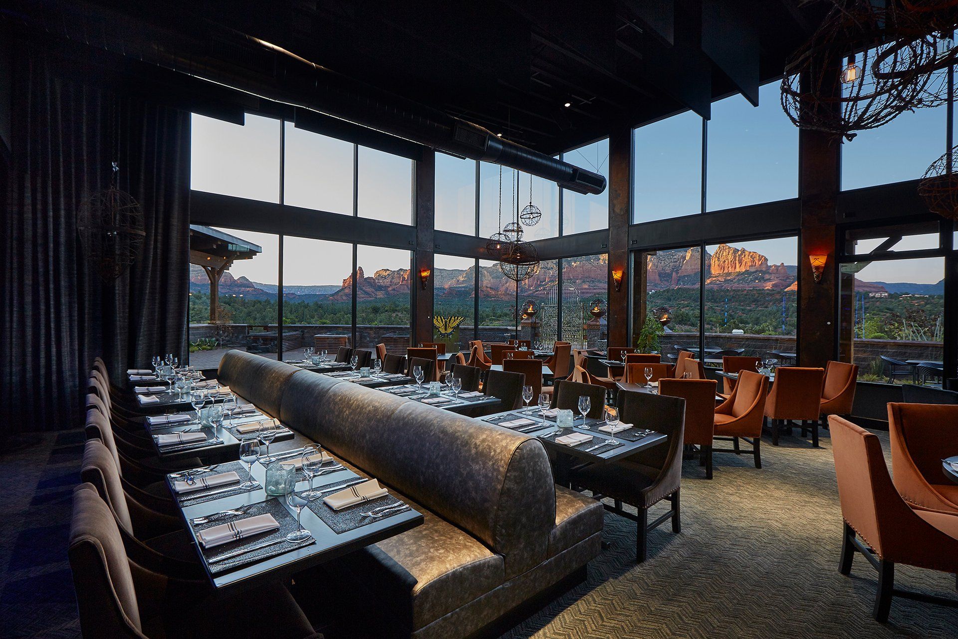 Restaurant interior with large windows overlooking desert landscape; tables set, chairs, and a long booth. Mariposa Sedona