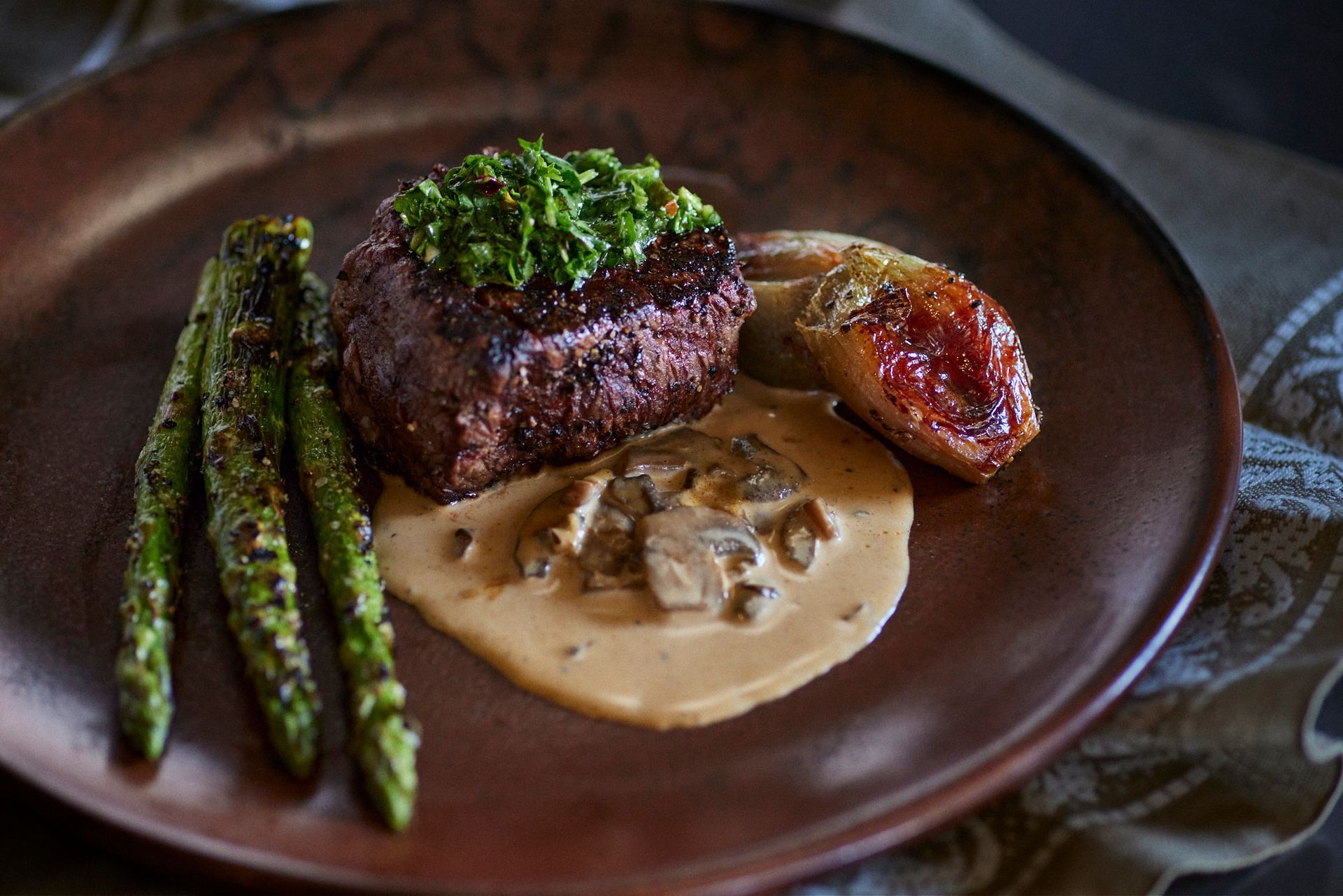 Steak topped with green sauce, mushroom sauce, asparagus, and a potato on a brown plate. Mariposa Sedona