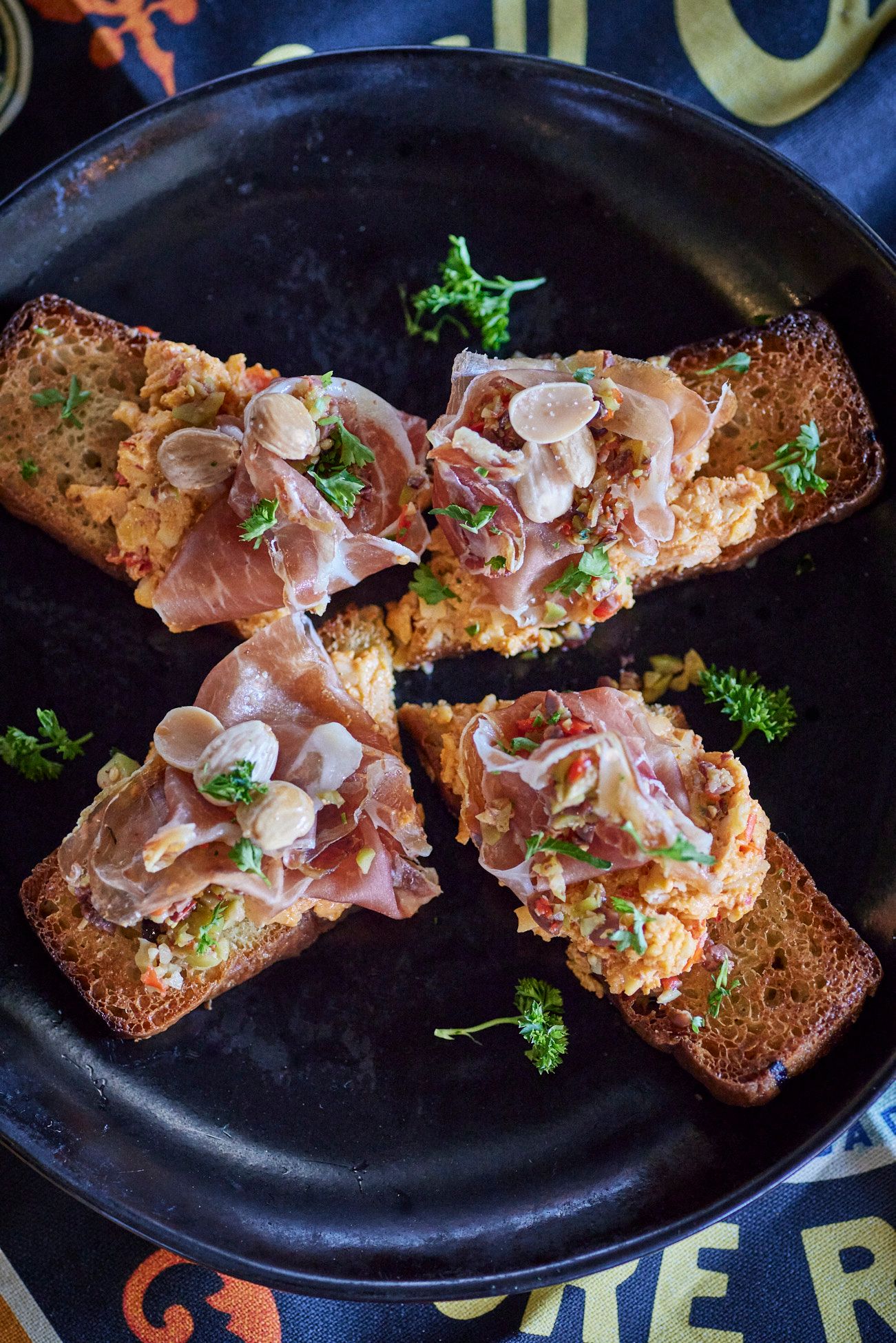 Toasted bread topped with orange spread, prosciutto, and nuts on a black plate. Mariposa Sedona