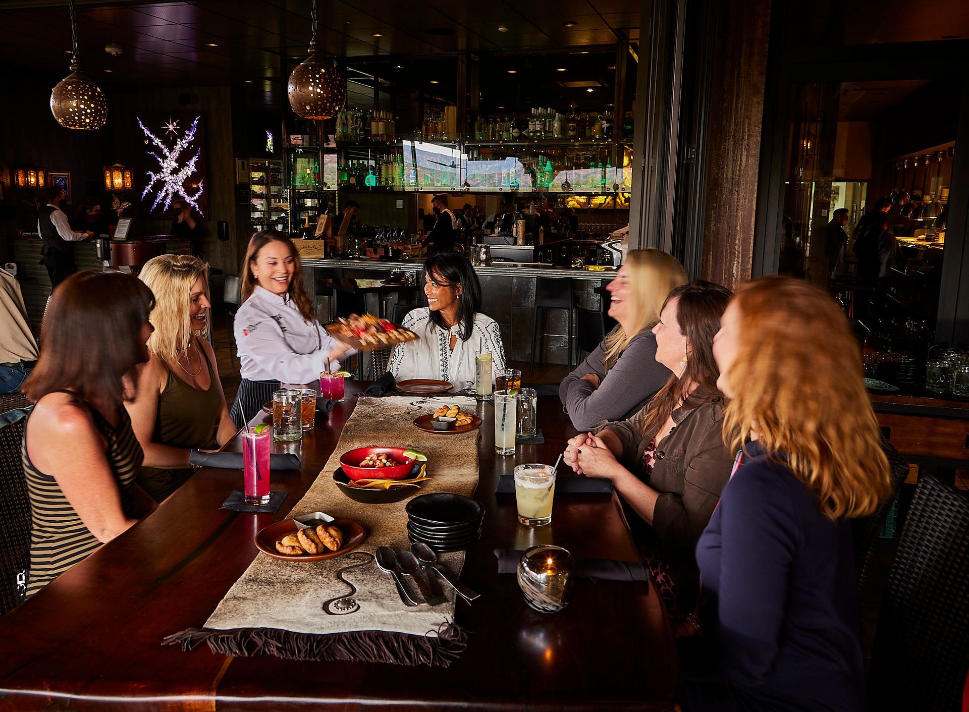 A group of women at a dimly lit bar Mariposa Sedona Bar, drinks and appetizers on the table, served by a smiling woman.
