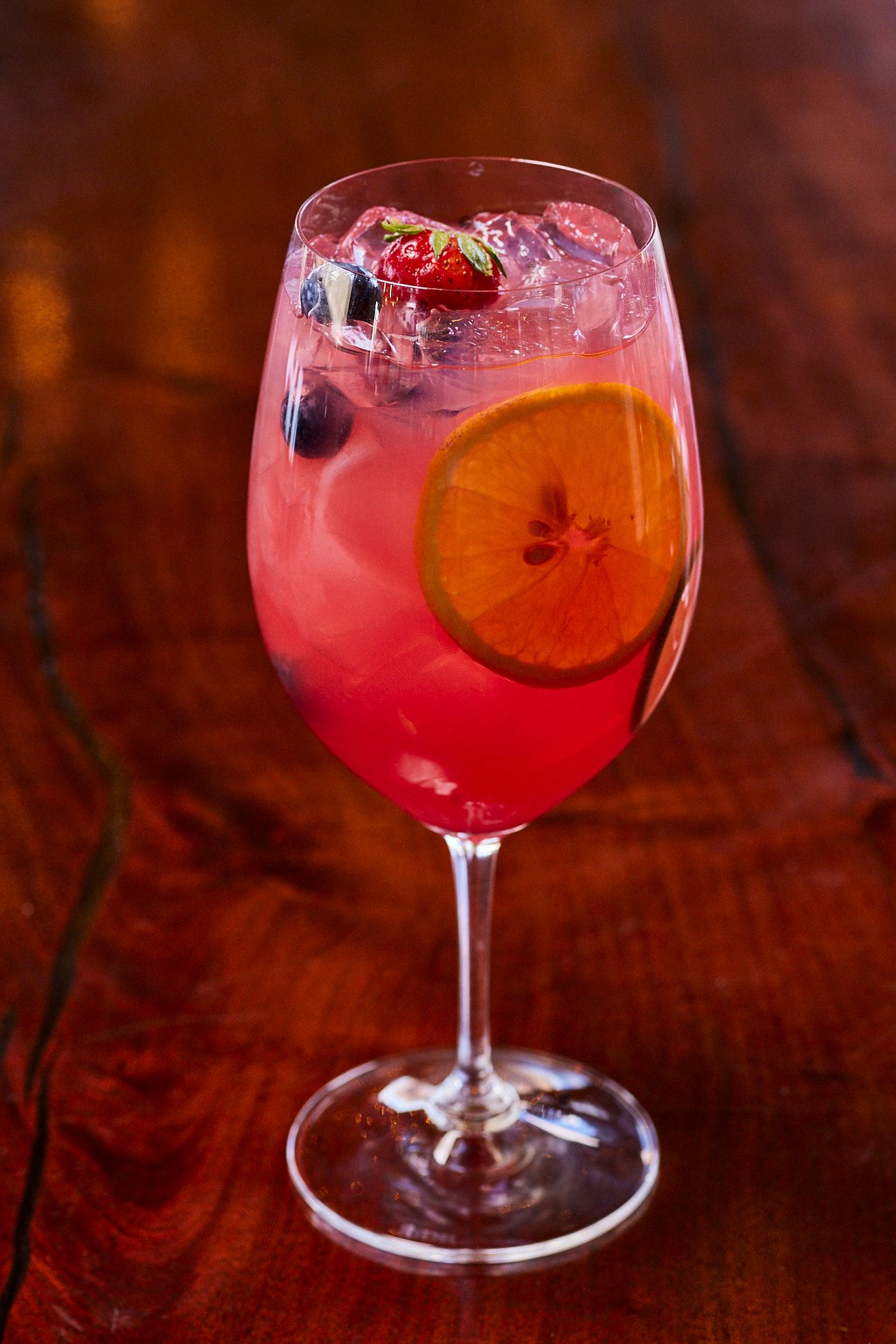 Pink cocktail with orange slice, blueberries, strawberry in a wine glass on a wooden table. Mariposa Sedona
