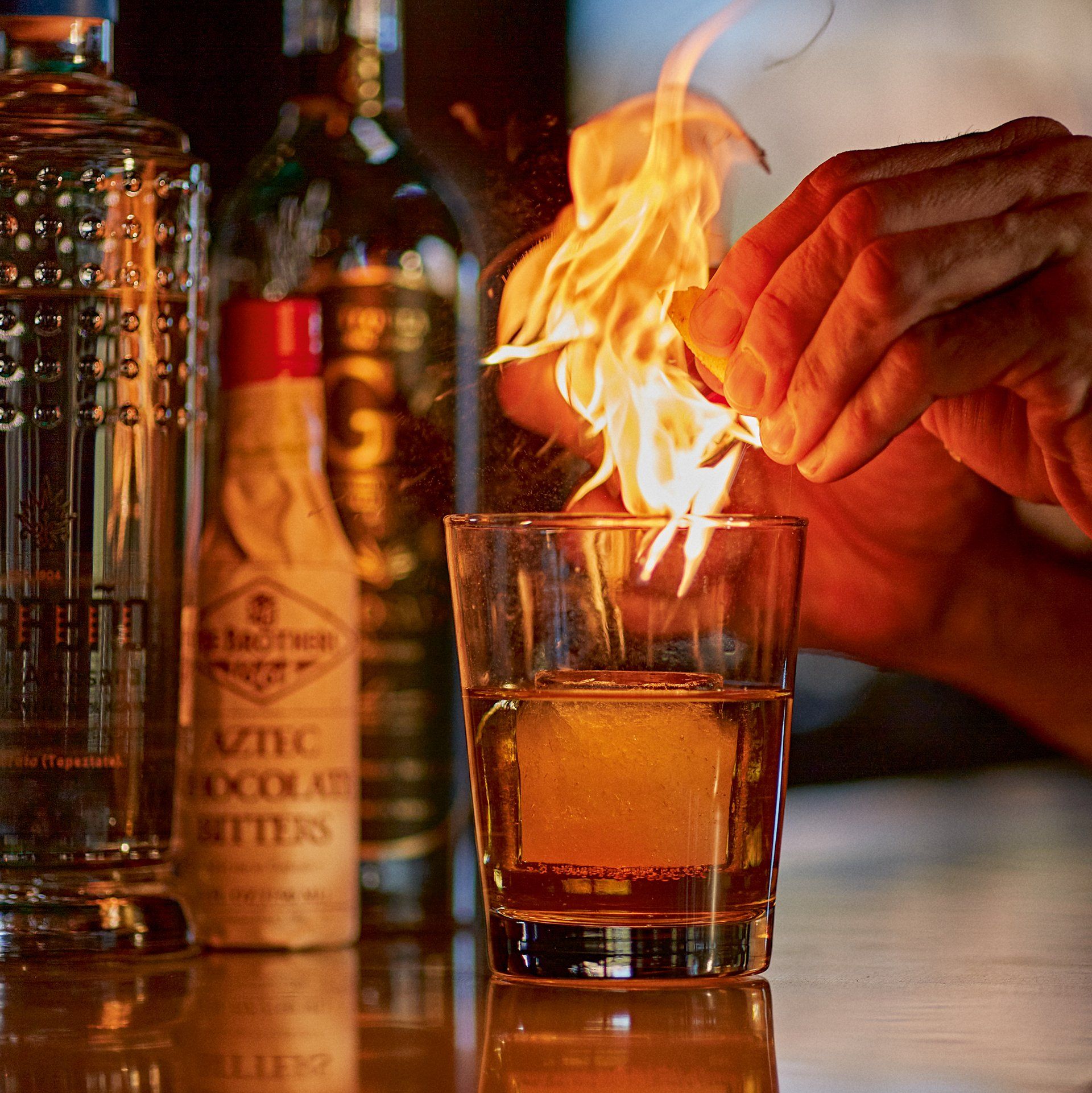 Hands lighting a flaming drink. Glass with ice, bottles of liquor behind. Mariposa Sedona