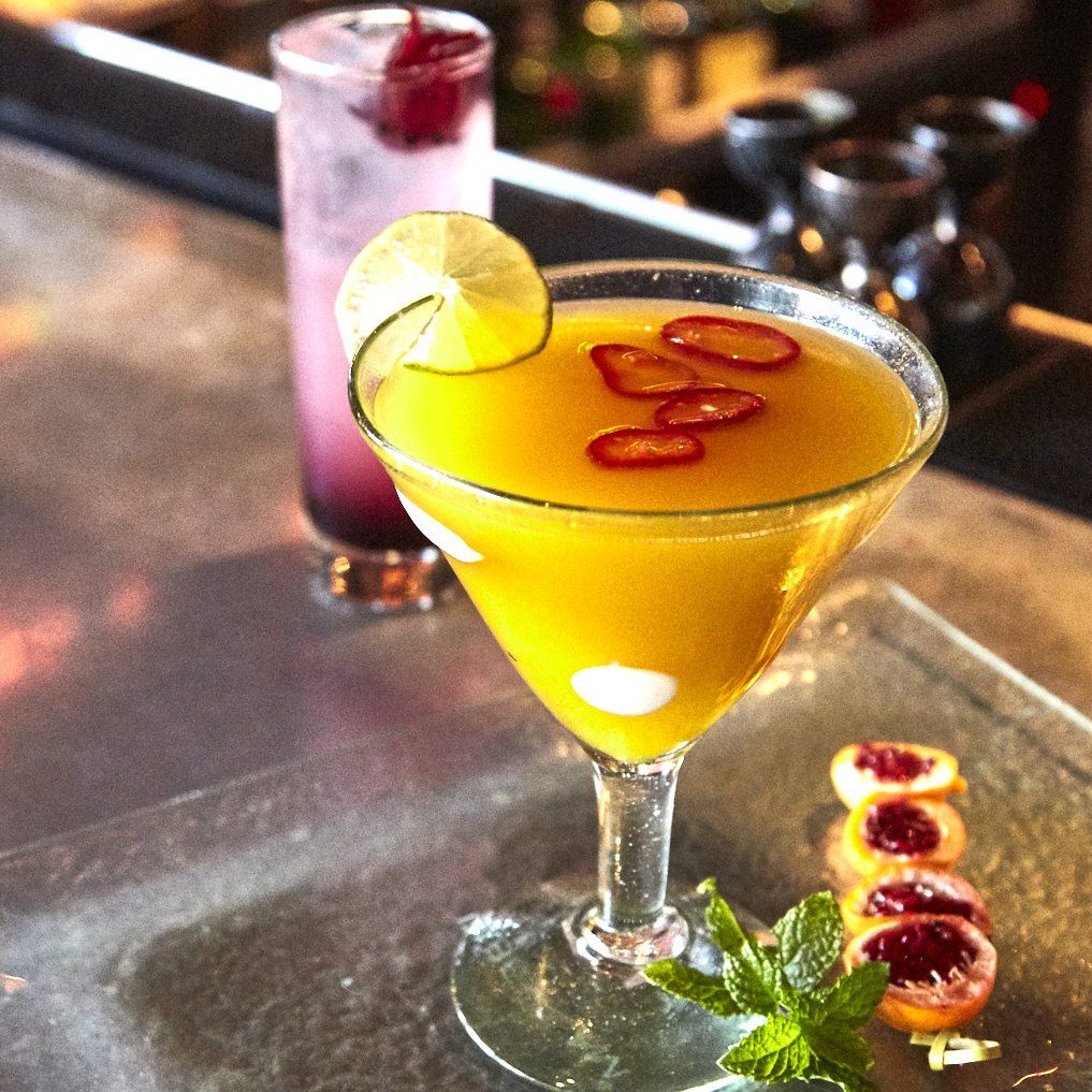 Yellow cocktail with lime and chili garnish on a glass plate; purple cocktail in the background. Mariposa Sedona