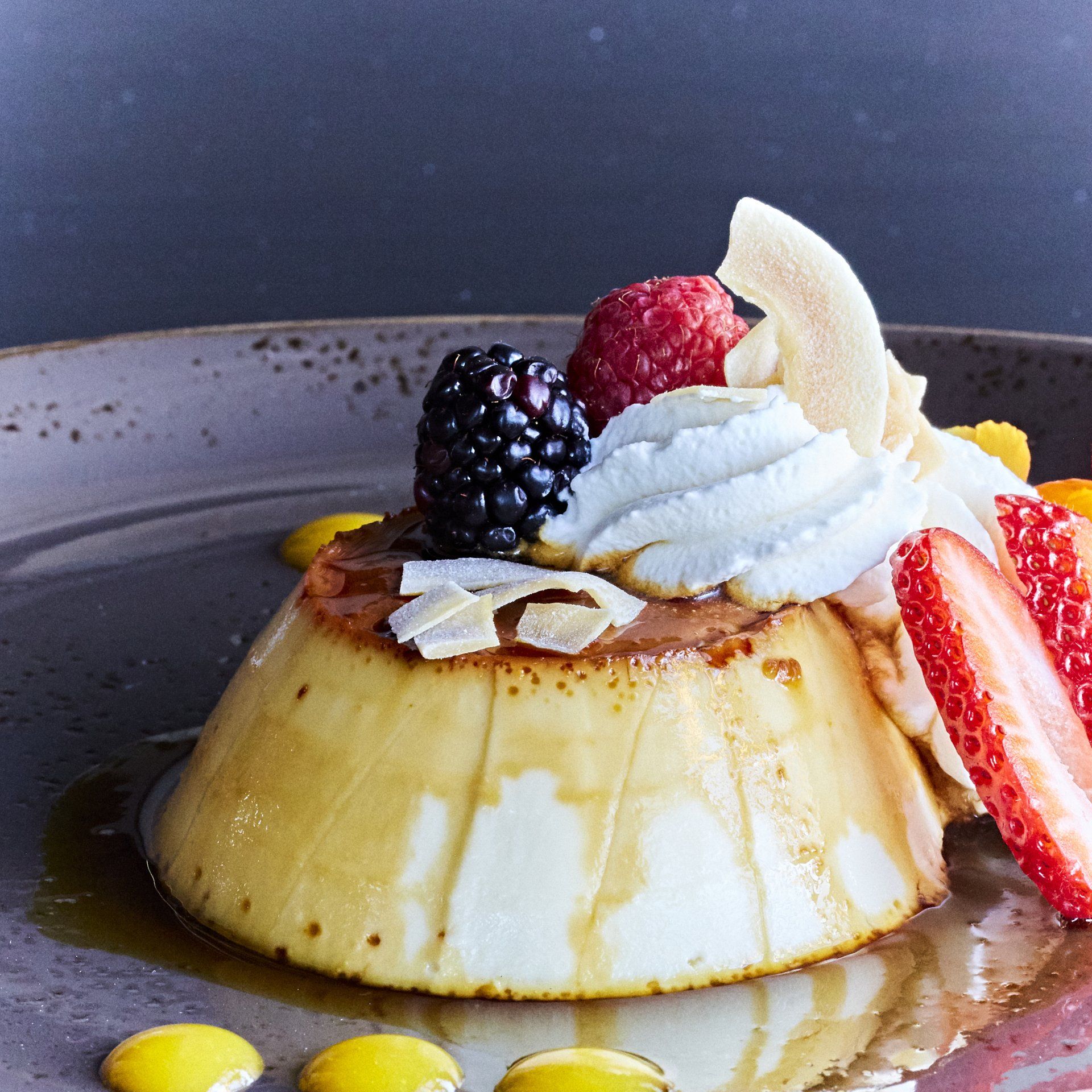 Flan dessert with whipped cream, berries, coconut flakes, and sliced strawberries on a grey plate.