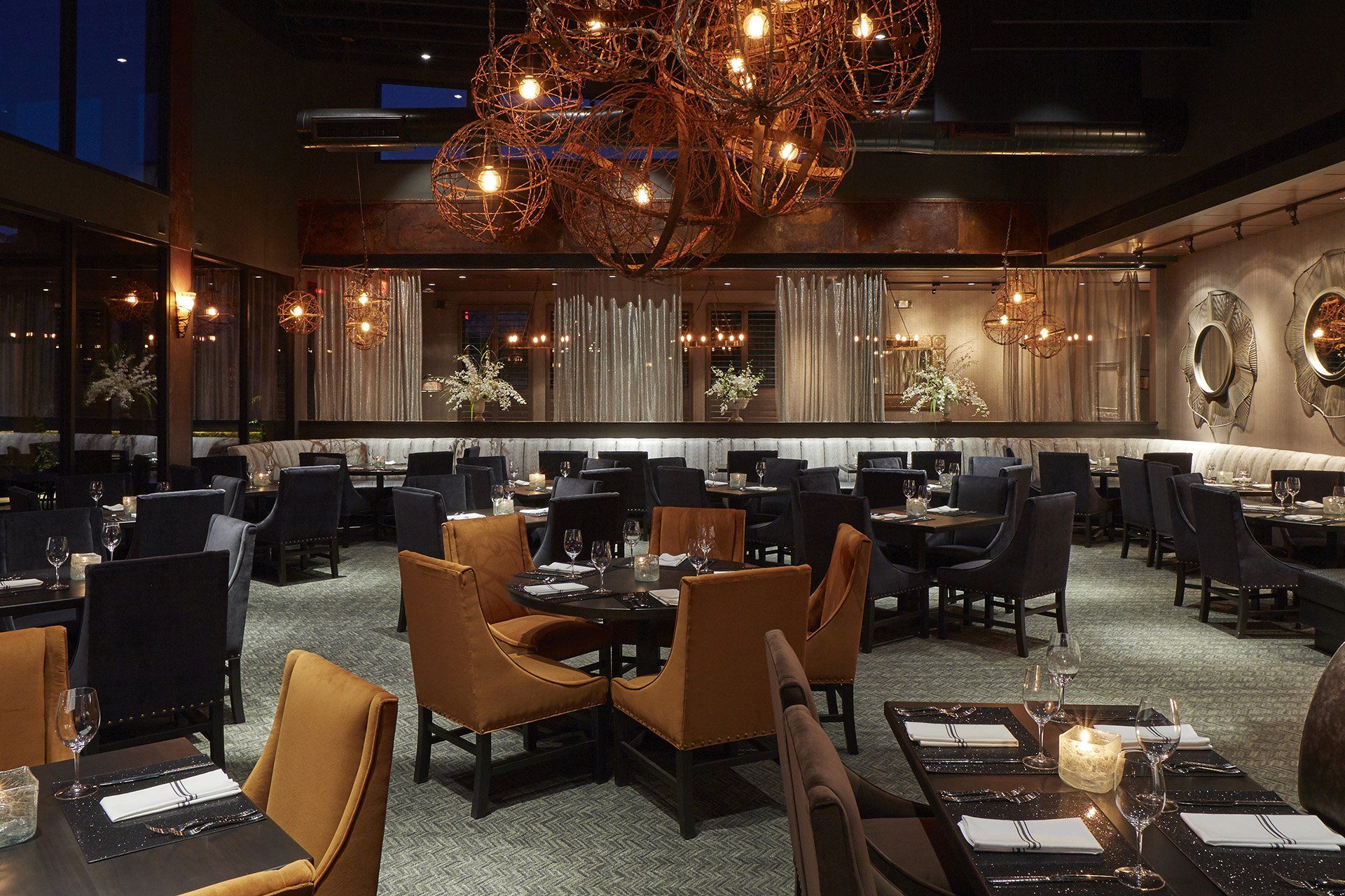 Upscale restaurant interior with dark tables and gold and dark blue chairs under artistic light fixtures.