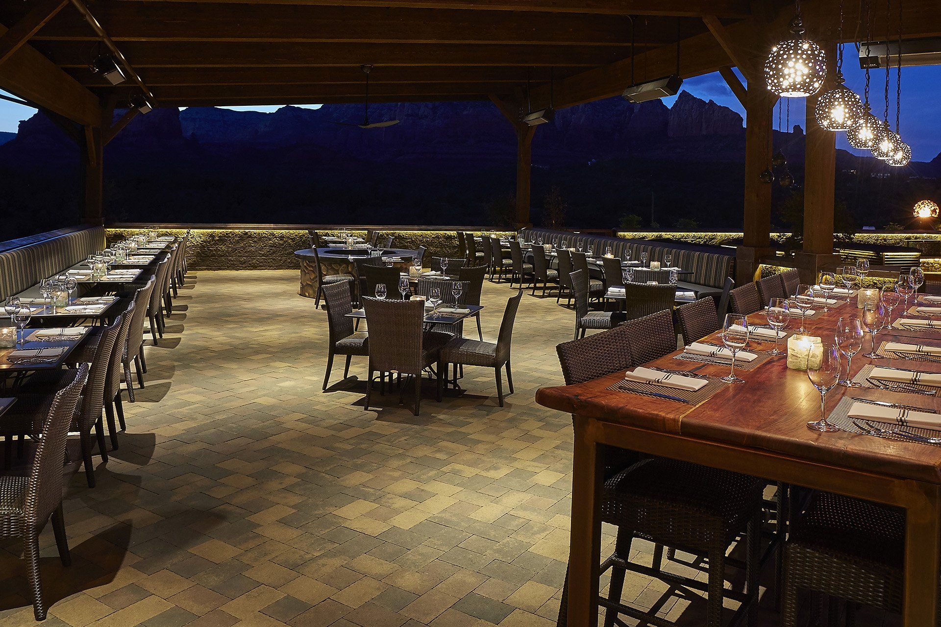 Outdoor restaurant patio at dusk with tables, chairs, and hanging lights, with mountains in the background. Mariposa Sedona