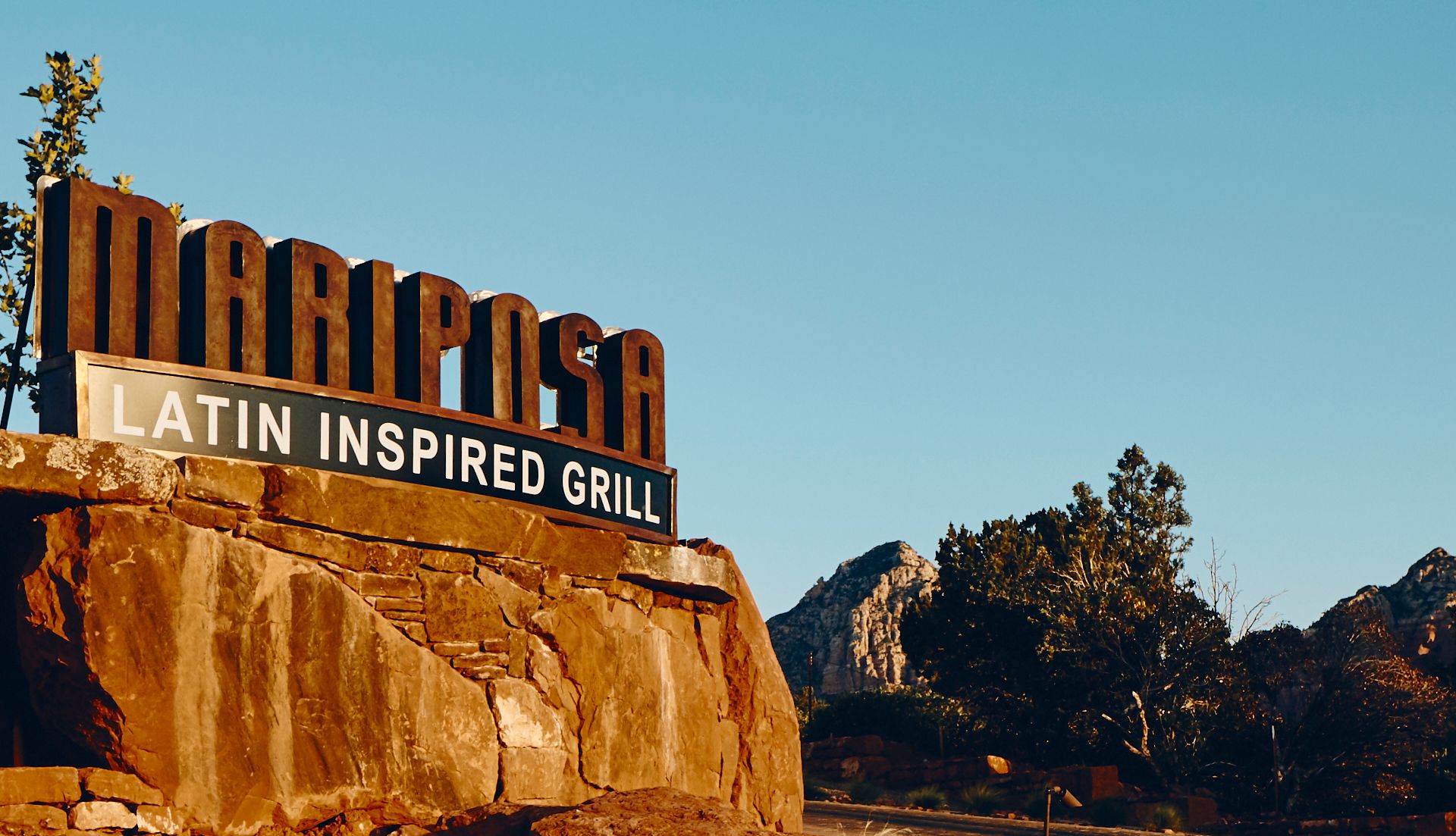Mariposa Latin Inspired Grill sign, brown rock wall, mountains, clear blue sky. Mariposa Sedona