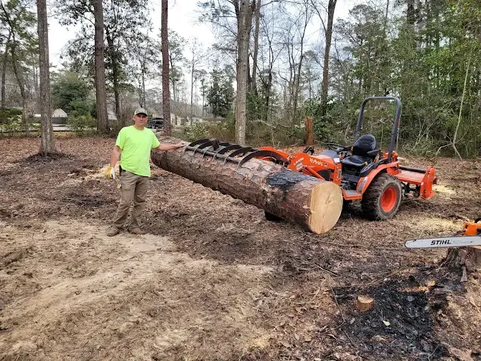 Man standing next to a large log held by a small orange tractor in a wooded area.